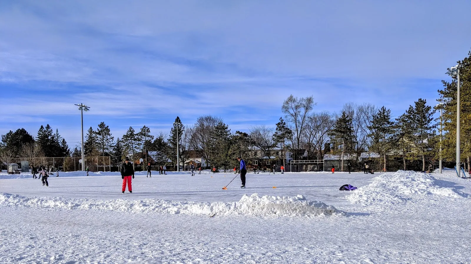 Patinadores y un jugador manejando el disco en la amplia pista de McKellar Park en un soleado día de invierno, con montones de nieve alrededor del hielo. — McKellar Park Outdoor Rink