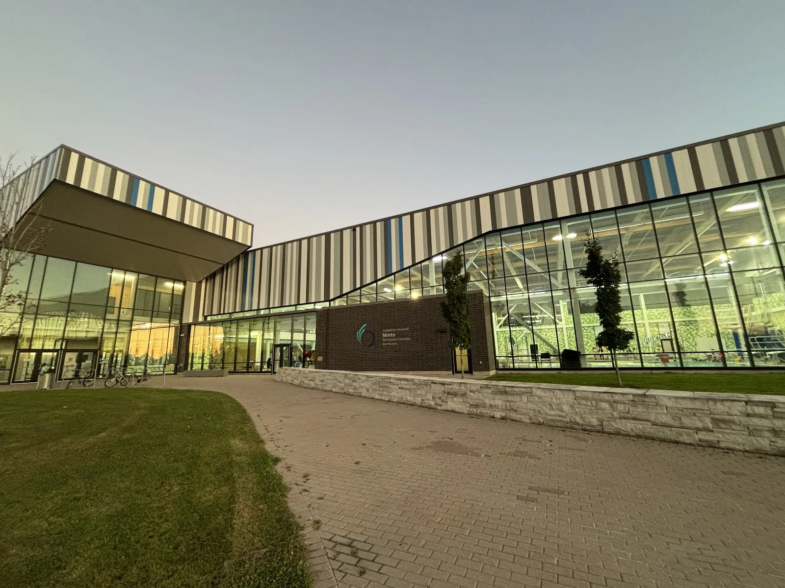 The glass-fronted Minto Recreation Complex in Barrhaven at dusk, with its striped metal cladding, the pool hall glowing behind the glazing, the name sign on the brick wall by the entrance and a paved walk and lawn in front. — Minto Recreation Complex – Barrhaven