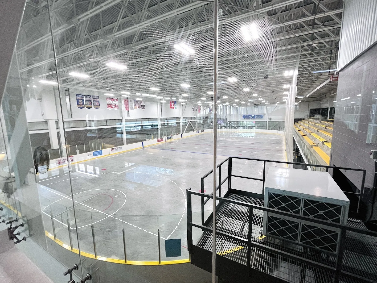 An empty rink at the Minto Recreation Complex seen from the upper level, with yellow bleachers and banners along the far wall. — Minto Recreation Complex – Barrhaven
