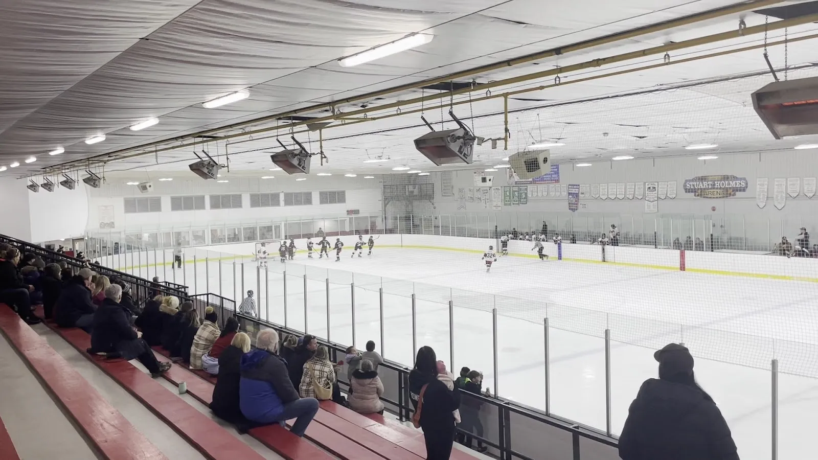 Spectators seated on red bleachers along the glass watching a game on a wide sheet, a long grey ceiling with ductwork and lights above the white ice. — Osgoode Community Centre and Stuart Holmes Arena