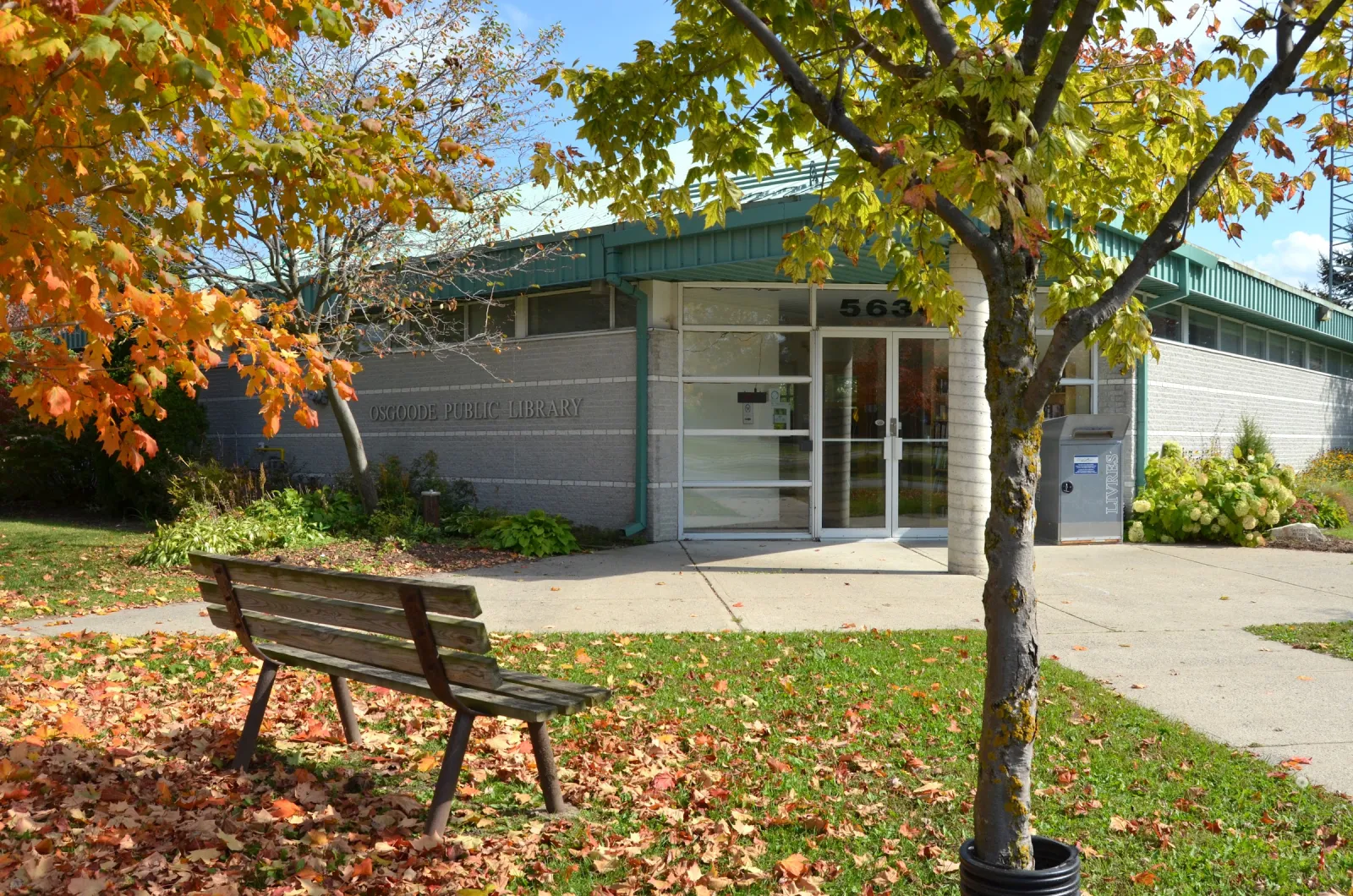 La entrada del centro comunitario de Osgoode en otoño, un edificio bajo con un vestíbulo de vidrio, una banca y un maple que deja caer hojas naranjas sobre el pasto. — Osgoode Community Centre Outdoor Rink