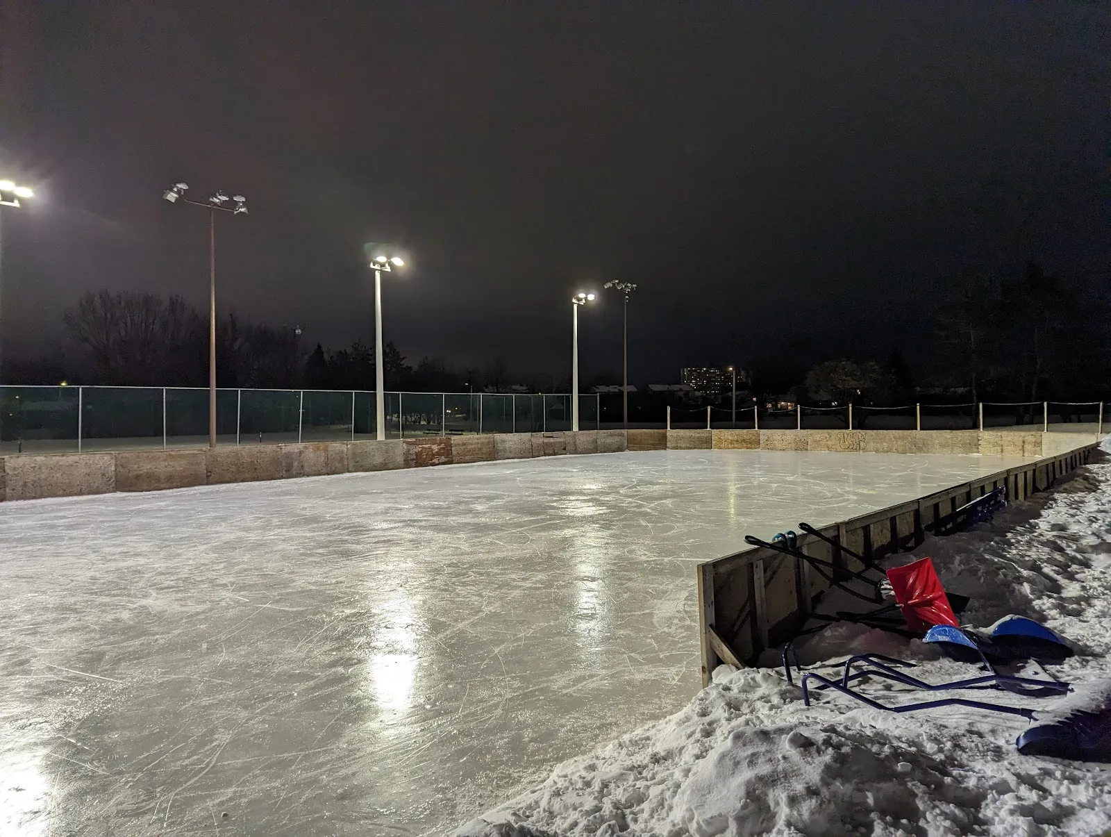 La pista exterior vacía con vallas en Owl Park de noche, con sus vallas color piedra, los reflectores y la cerca del lado opuesto, y un banco de nieve y palos de hockey a la derecha — Owl Park Outdoor Rink