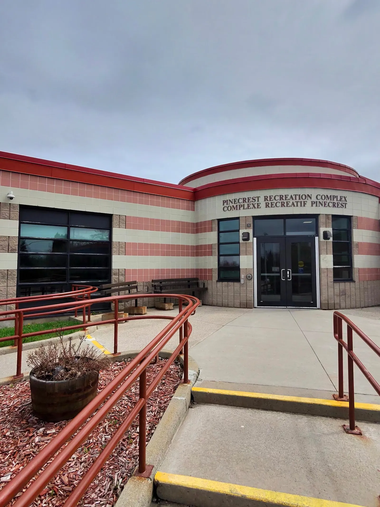 The entrance of the brick-striped building under the Pinecrest Recreation Complex / Complexe récréatif Pinecrest sign, with a ramp and red railings in front. — Pinecrest Recreation Complex – Barbara Anne Scott Arena