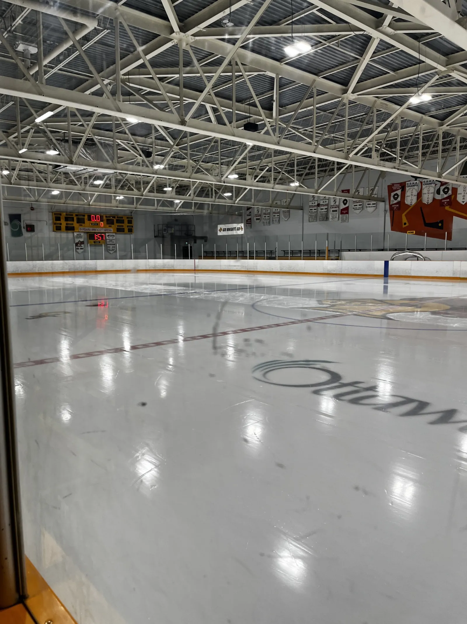 The empty ice seen through the glass, with the City of Ottawa logo at centre ice, the scoreboard and banners under a steel-truss roof. — Pinecrest Recreation Complex – Barbara Anne Scott Arena