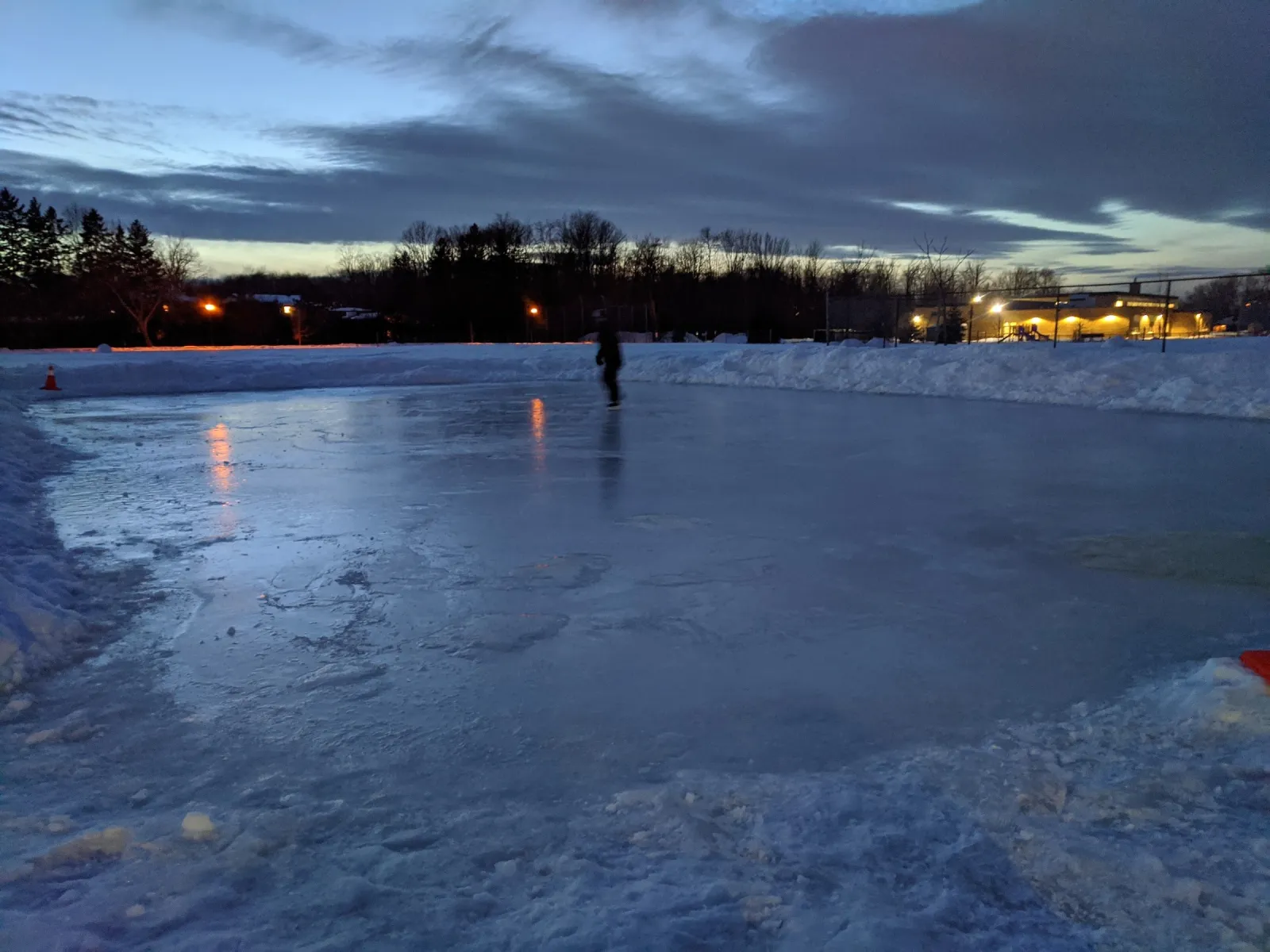 La pista al aire libre de Playfair Park al anochecer, con un patinador solitario en el hielo, montones de nieve alrededor y luces que brillan más allá de los árboles. — Playfair Park Outdoor Rink