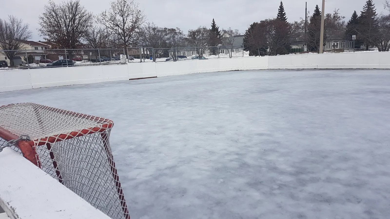 La pista al aire libre de Queenswood Ridge en invierno, una pista de hielo espolvoreada de nieve dentro de vallas blancas con una portería de hockey en la esquina. — Queenswood Ridge Park Outdoor Rink