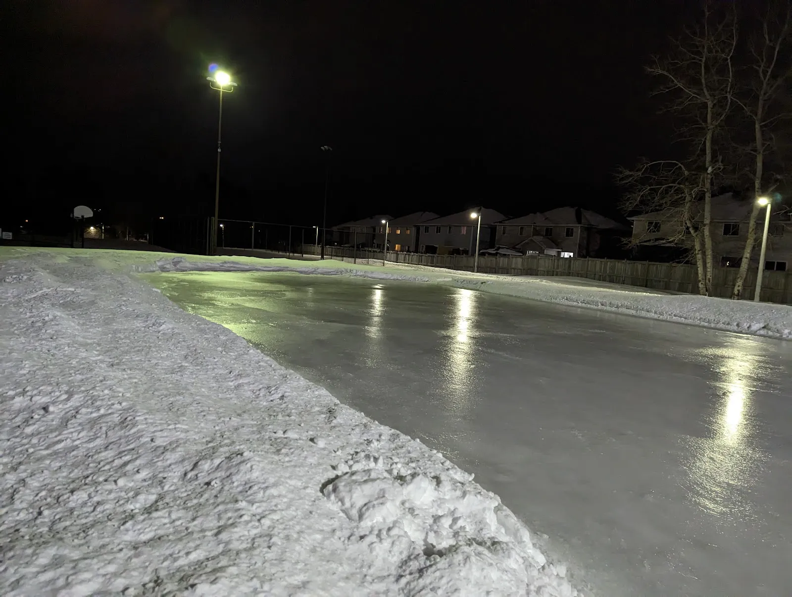 Smooth ice at the Ruth Wildgen Park rink at night under a floodlight, with a snow bank along the near edge and the houses behind the fence — Ruth Wildgen Park Outdoor Rink
