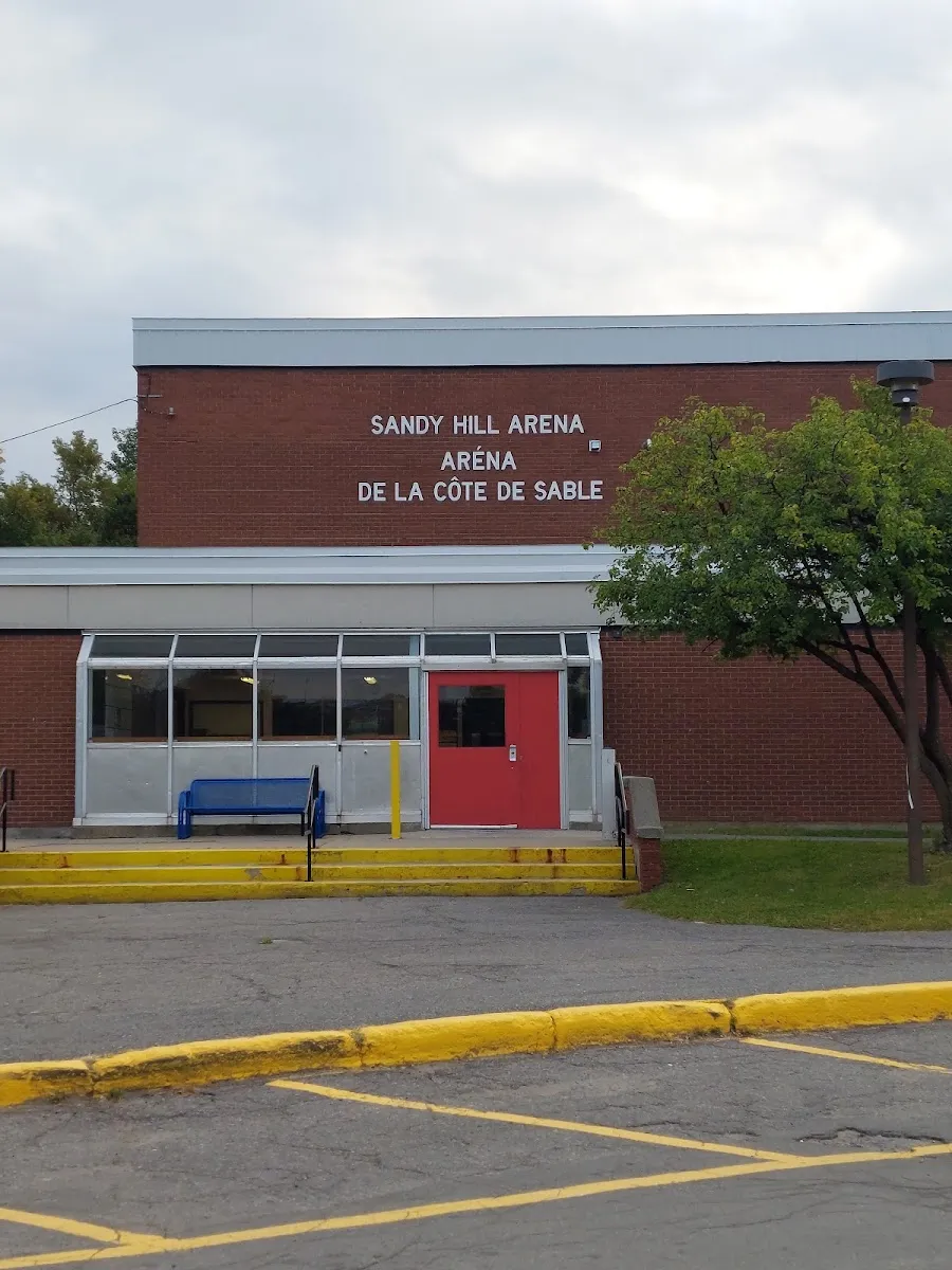 The Sandy Hill Arena entrance, its bilingual name on the brick wall above a red door and a blue bench. — Sandy Hill Arena