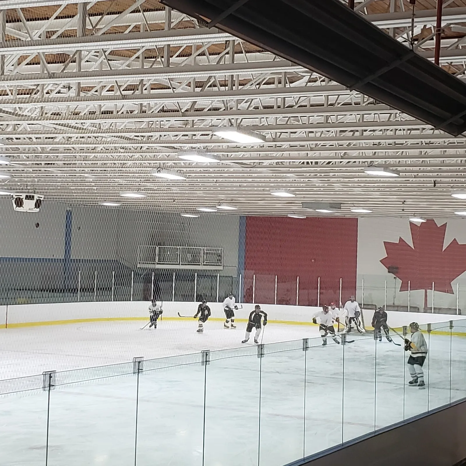 A game under a steel-trussed roof with a Canadian maple leaf on the red panel behind, players spread across the far end of the sheet. — Sandy Hill Arena