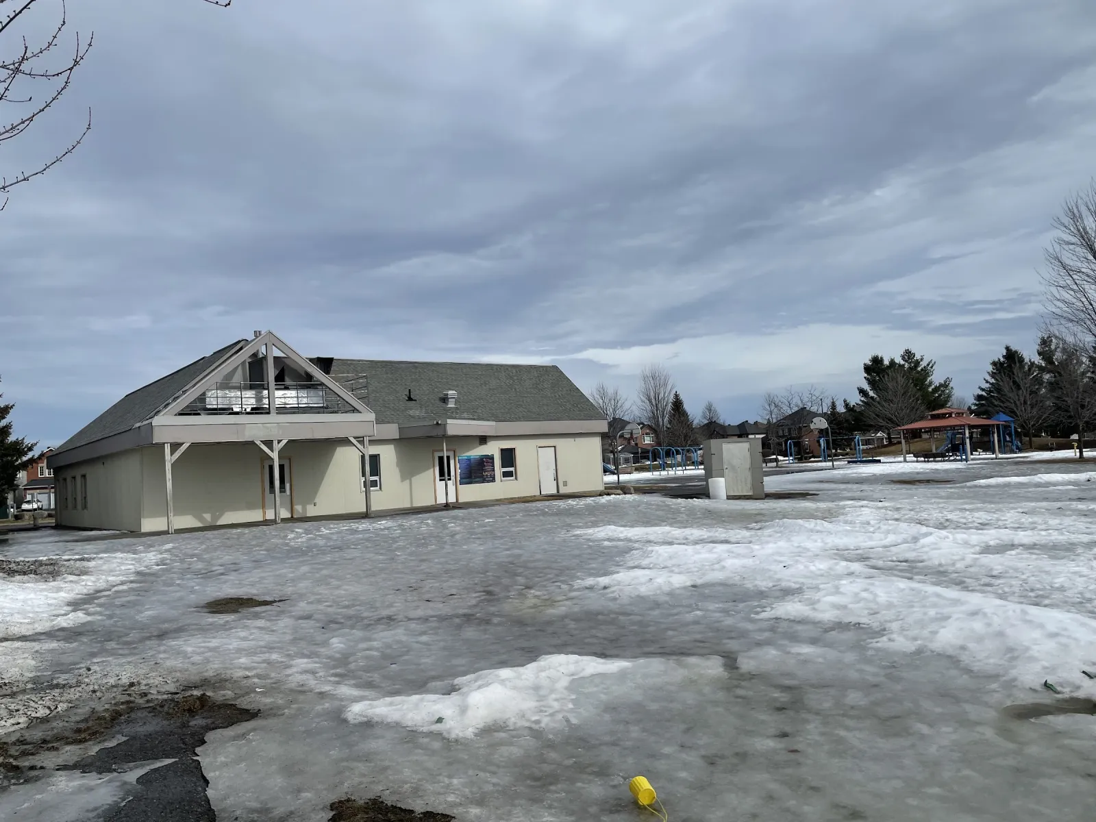 The Stonecrest Park field house, a pale clapboard building with a gabled roof, across a lawn of melting snow in early spring. — Stonecrest Park Outdoor Rink