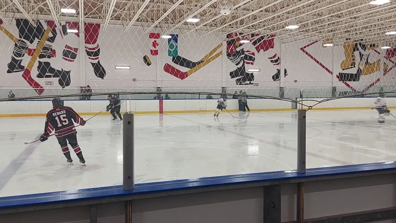Players spread across a wide sheet beneath a long white wall painted with hockey figures, seen over the glass. — Tom Brown Arena