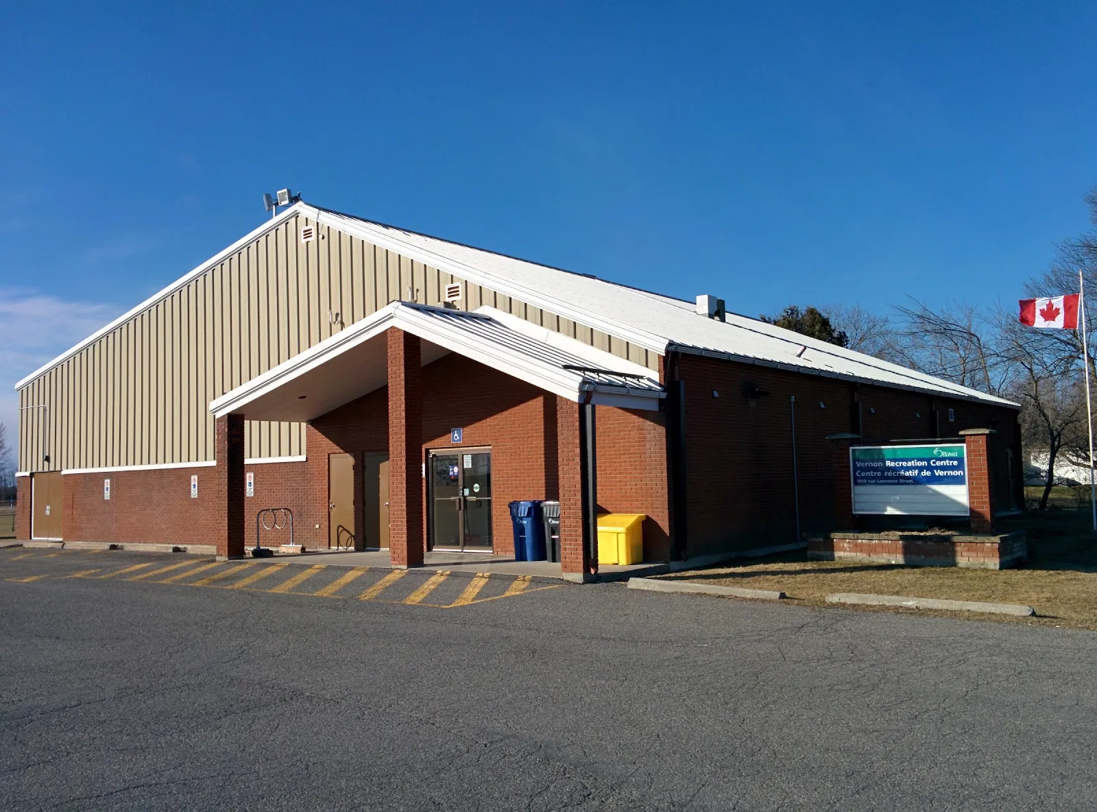 El Vernon Recreation Centre en un día soleado: un salón de ladrillo con una sección alta con revestimiento metálico, una entrada techada y el letrero del centro al frente. — Vernon Recreation Centre Outdoor Rink