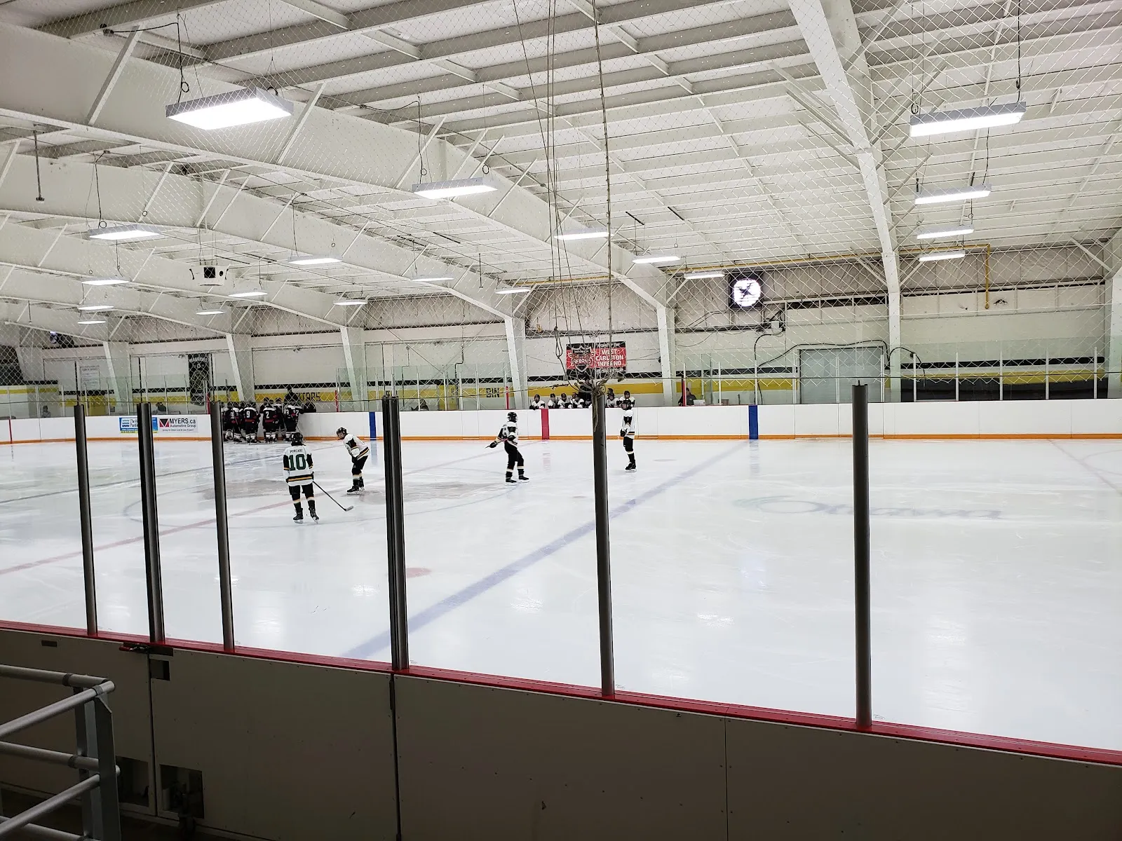 Full ice sheet during a game, shot from behind the glass, with a West Carleton Minor Hockey banner and a Myers.ca Automotive Group ad board on the boards. — W. Erskine Johnston Arena