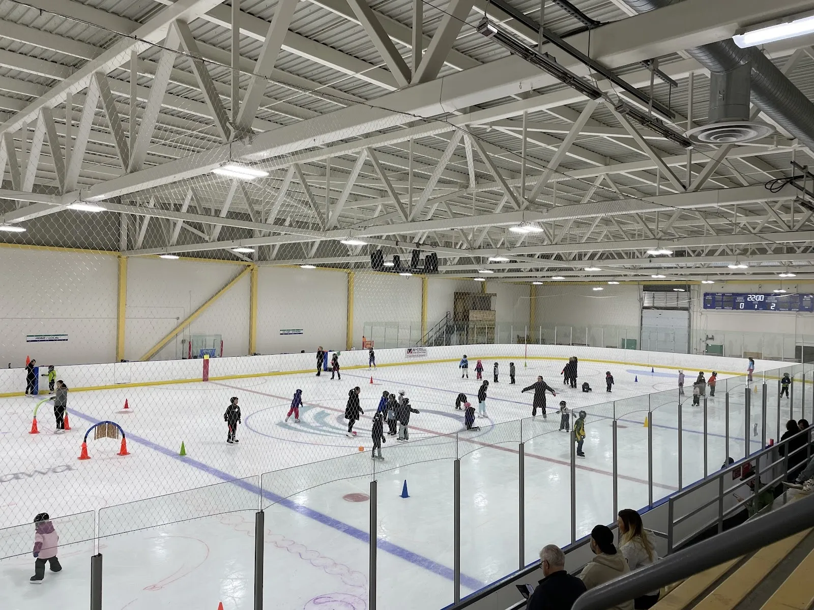 Una sesión pública en una pista amplia vista desde las gradas, con patinadores repartidos entre conos y una esquina con línea azul bajo un techo de armaduras de acero. — Walter Baker Sports Centre