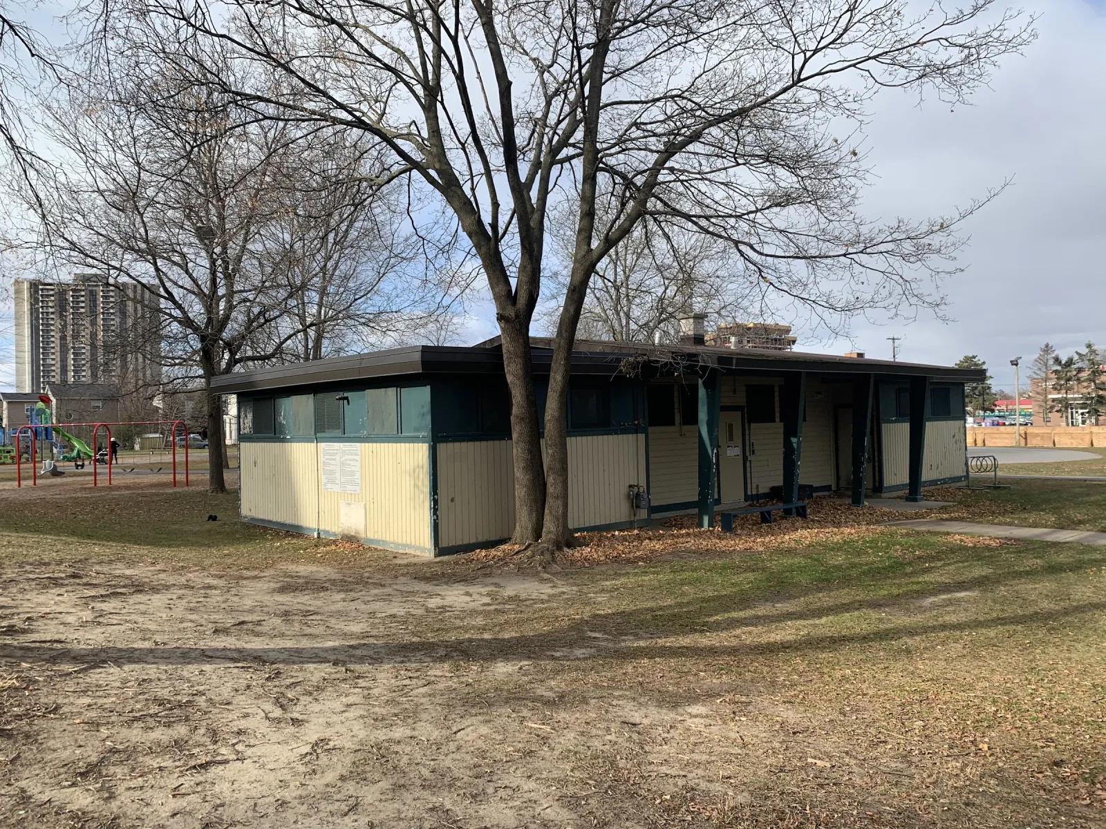 La caseta de Woodroffe Park, un edificio bajo verde y crema bajo árboles sin hojas, con el área de juegos y edificios de departamentos más allá. — Woodroffe Park Outdoor Rink