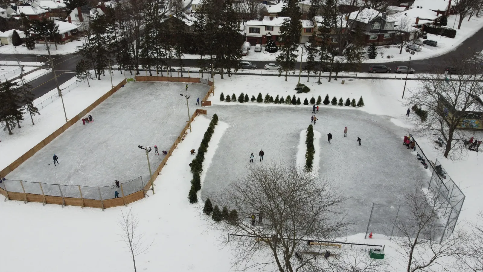 Woodroffe Park desde arriba en invierno: la pista de hockey con vallas junto a una superficie más grande de patinaje recreativo, ambas con patinadores, entre pastos nevados y casas. — Woodroffe Park Outdoor Rink