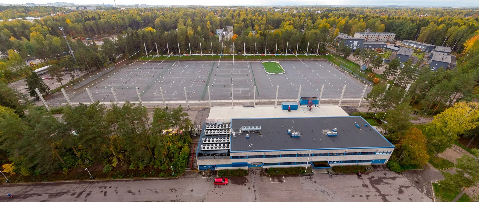 Aerial view of the Oulunkylä artificial ice rink in Helsinki in autumn, with the service building in front and trees around the rink. — Oulunkylän tekojääkenttä