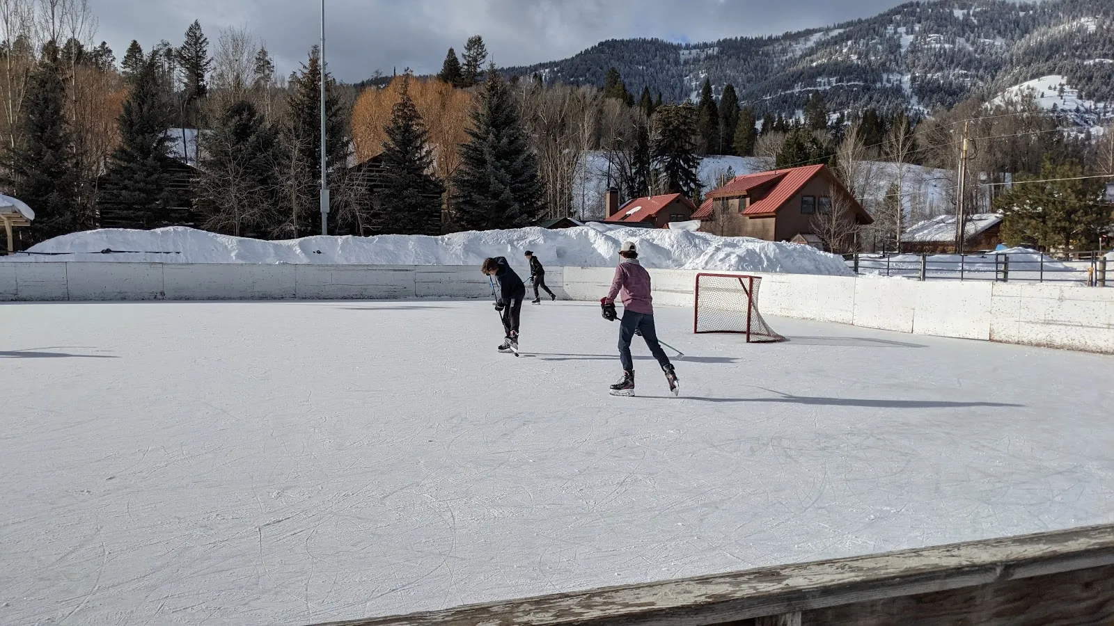 Two skaters on the rink's ice in daylight, a hockey net and mountains behind — Owen Bircher Park Rink