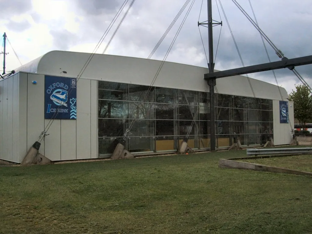 The low silver-grey Oxford Ice Rink building seen across grass, with a blue 'Oxford Ice Rink' sign on the end wall, glazed side wall and the mast-and-cable roof supports above. — Oxford Ice Rink
