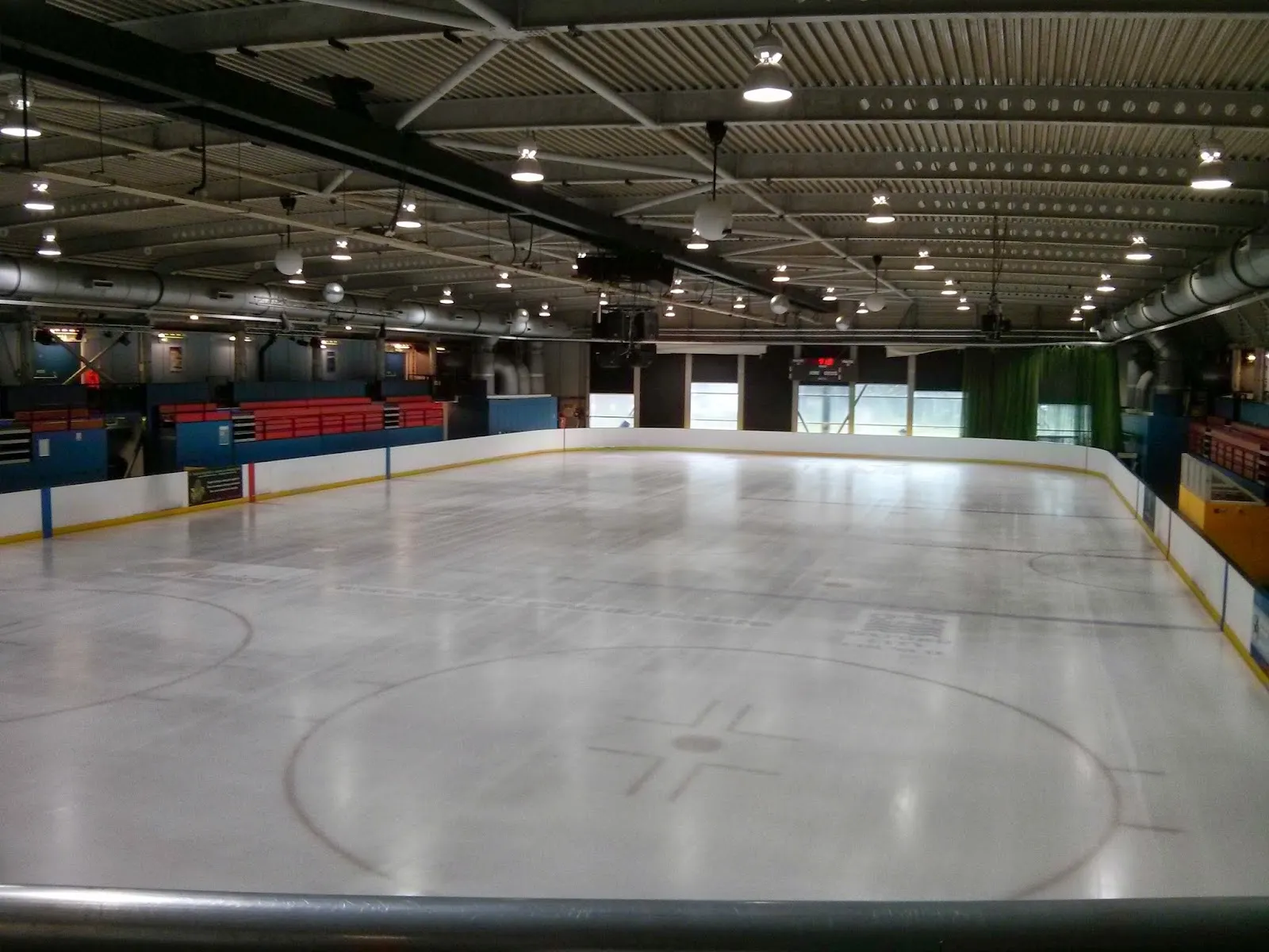 The empty ice sheet from the rail, with the blue and red boards, the stands and the dark steel-beamed roof behind. — Oxford Ice Rink