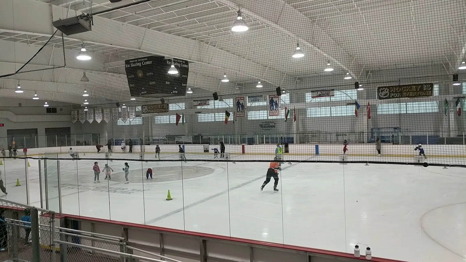Una sesión pública en la pista principal del centro vista sobre las bandas: patinadores dispersos y conos naranjas, el marcador del Town of Oyster Bay Ice Skating Center y banderines colgados bajo el techo a la vista, con red de protección a lo largo del vidrio. — Town of Oyster Bay Ice Skating Center