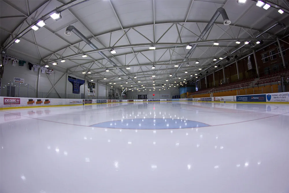 The empty white ice sheet of the OZO Halle seen at low angle, with sponsor boards along the left, blue centre circle, a steel-truss roof with floodlights and red tiered seating on the right. — OZO Halle