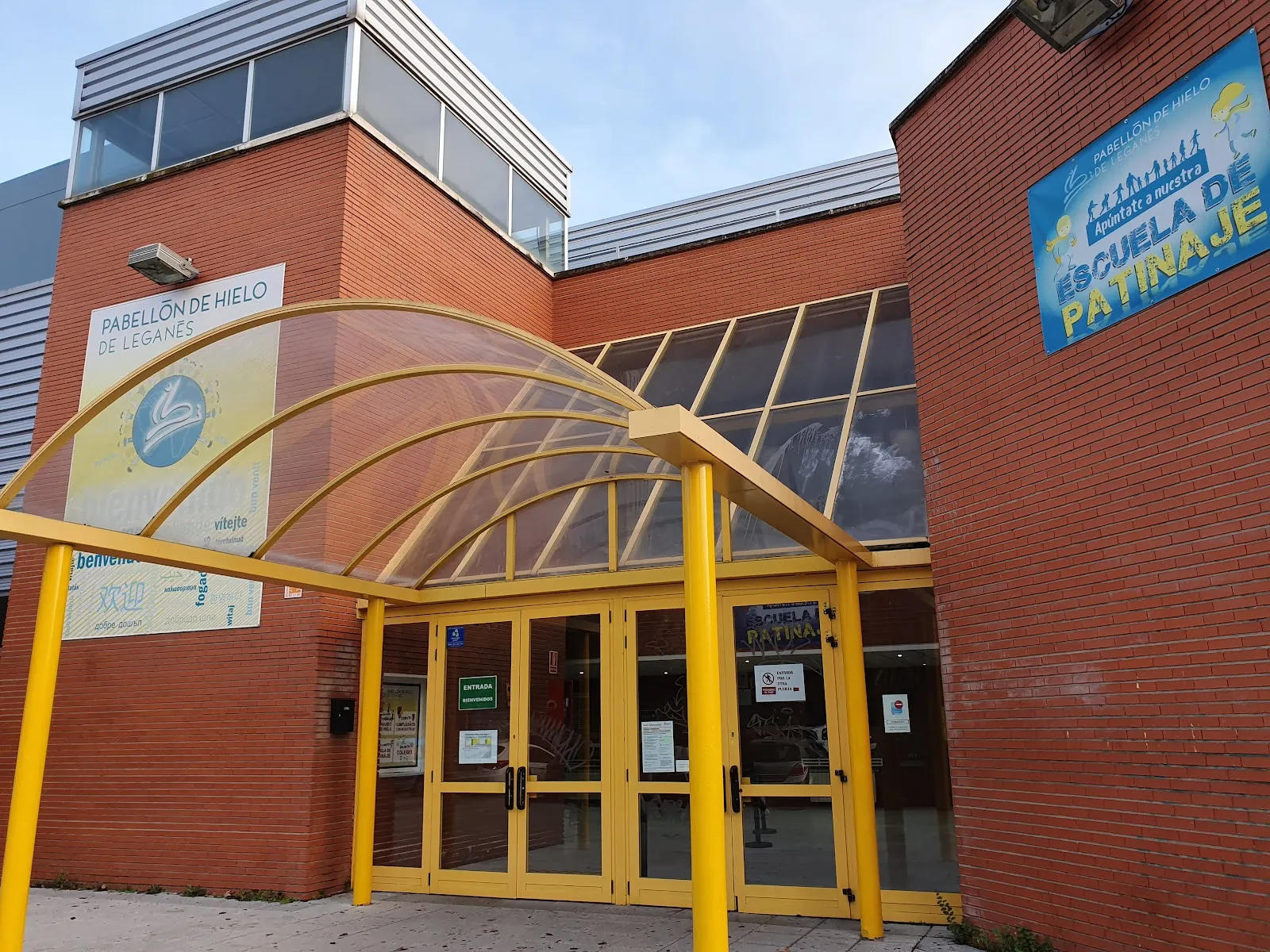 The red-brick entrance of the Pabellón de Hielo de Leganés under its curved yellow canopy, with the pavilion's name banner and the skating school sign — Pabellón de Hielo de Leganés