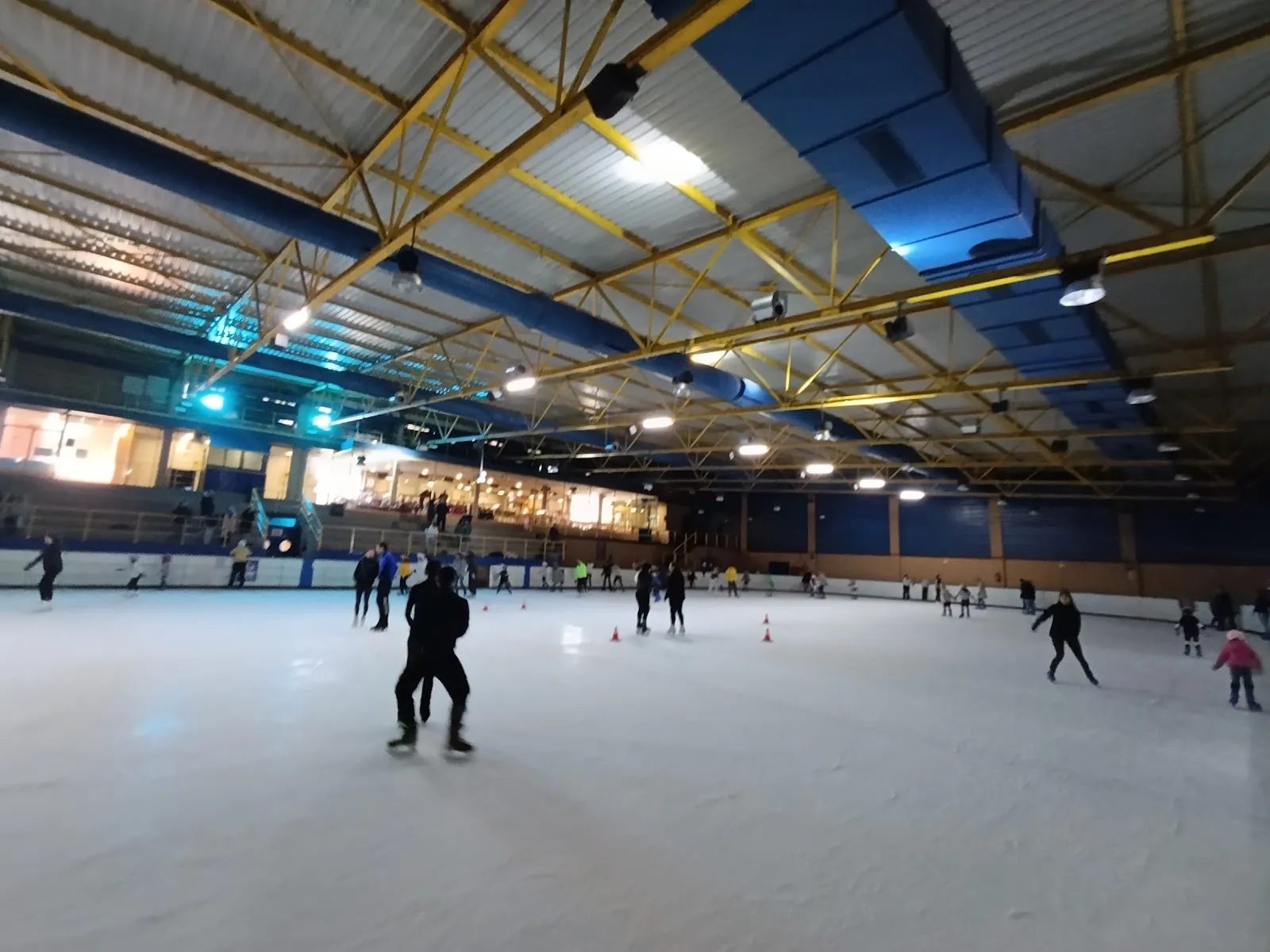 Wide view of the ice sheet in the Leganés ice pavilion, with a few skaters on the ice, the yellow steel roof trusses and the long side boards. — Pabellón de Hielo de Leganés
