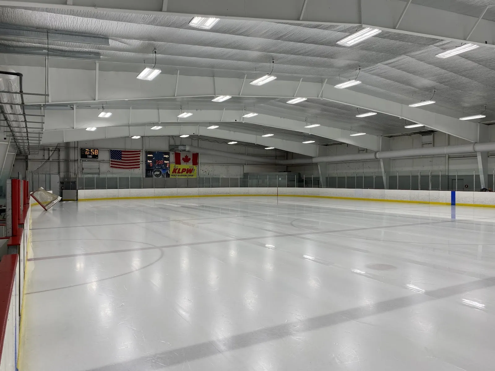 The empty ice sheet at Pacific Ice Rink with the boards, scoreboard and flags at the far end — Pacific Ice Rink
