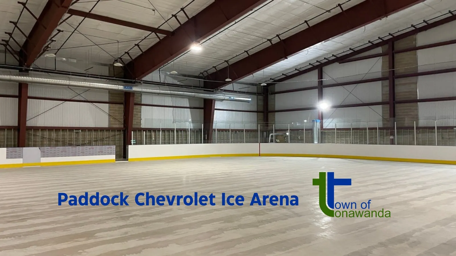 The empty Paddock Chevrolet Ice Arena ice with the arena name on the boards and steel roof trusses above. — Paddock Chevrolet Ice Arena