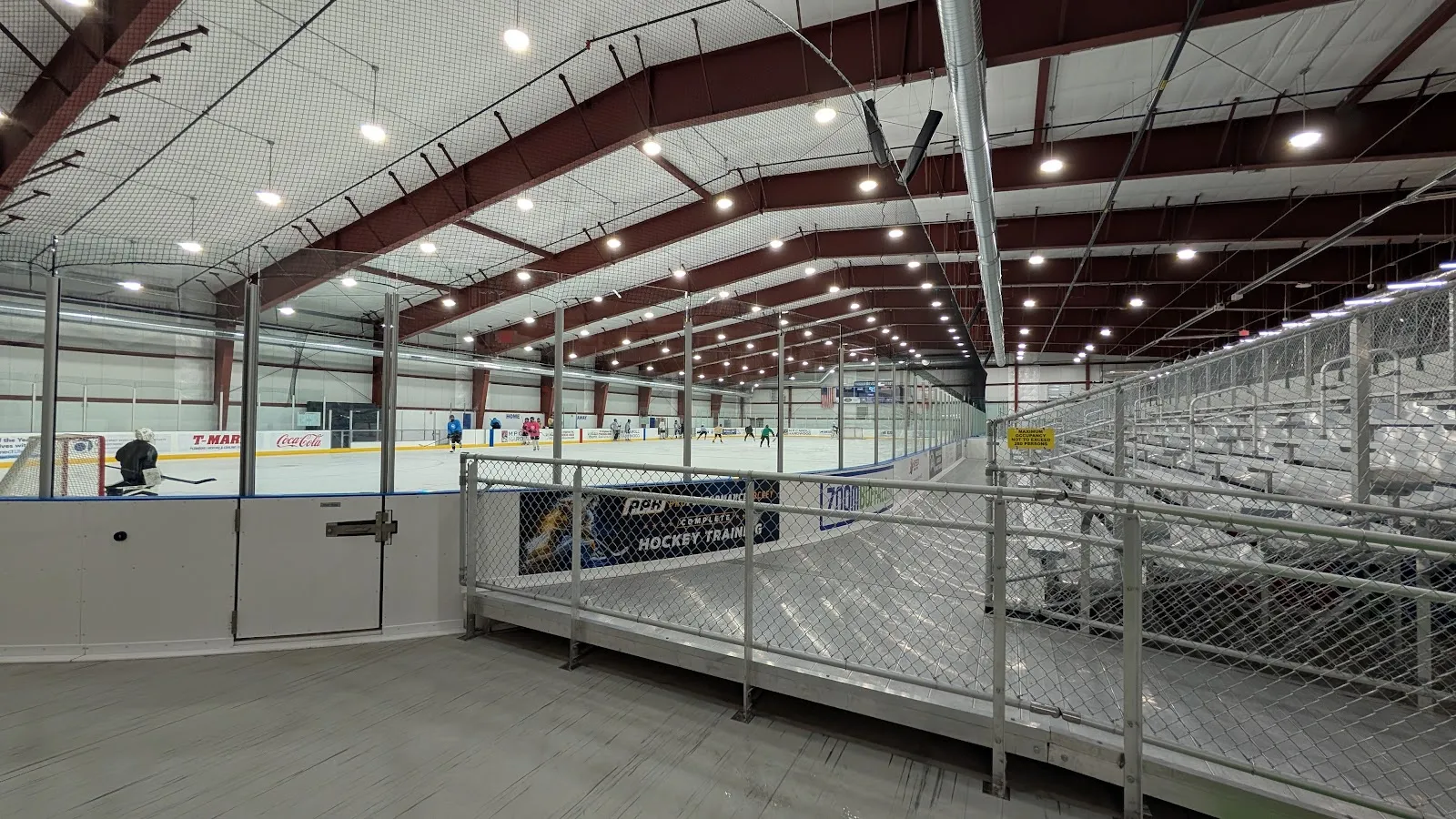 The Paddock Chevrolet Ice Arena rink seen from the stands, with a metal rail and steel roof structure overhead. — Paddock Chevrolet Ice Arena