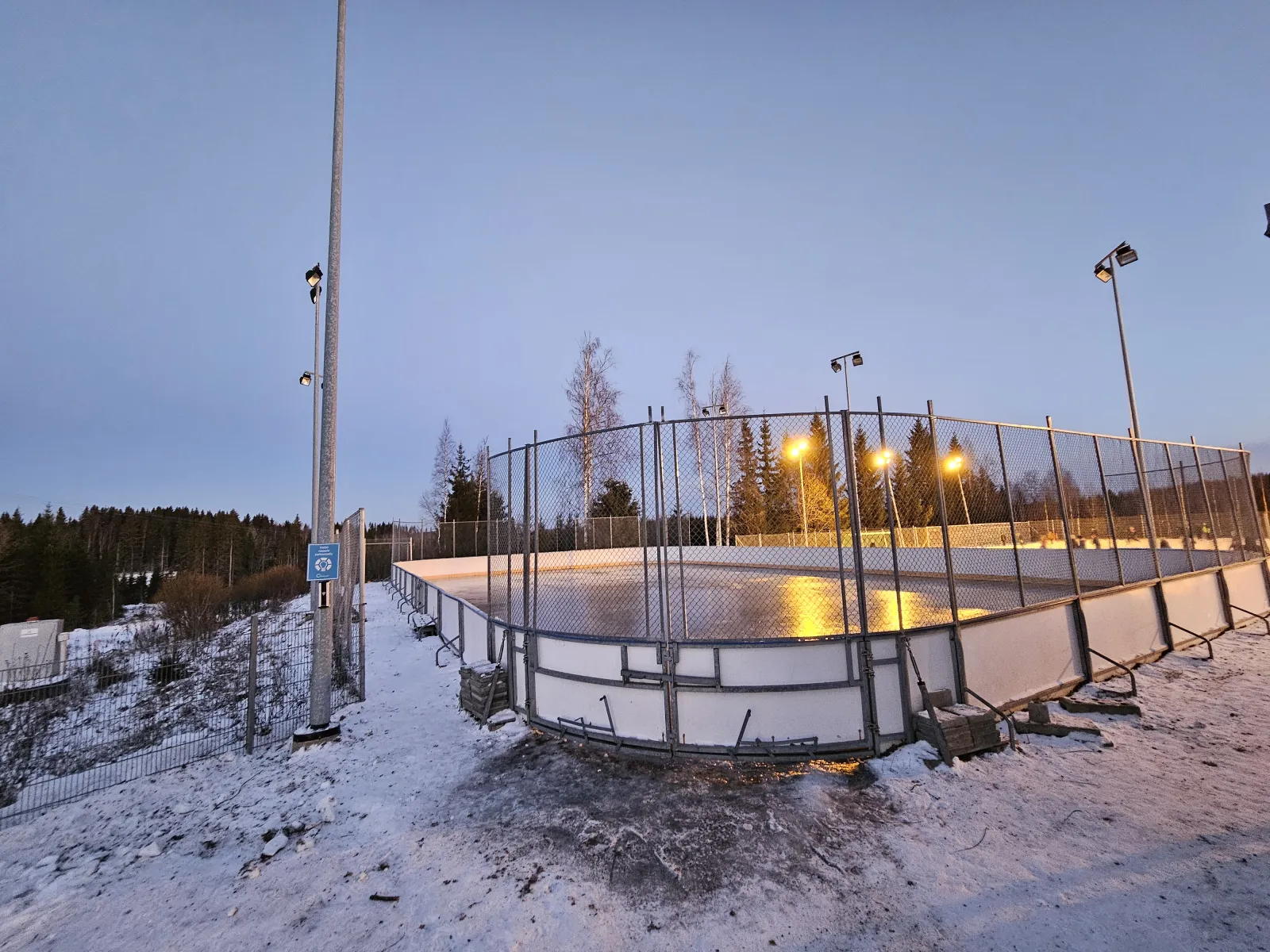 The boarded Pajulahti rink at dusk, lit by its floodlights, with snow around the boards. — Pajulahden koulun kaukalo