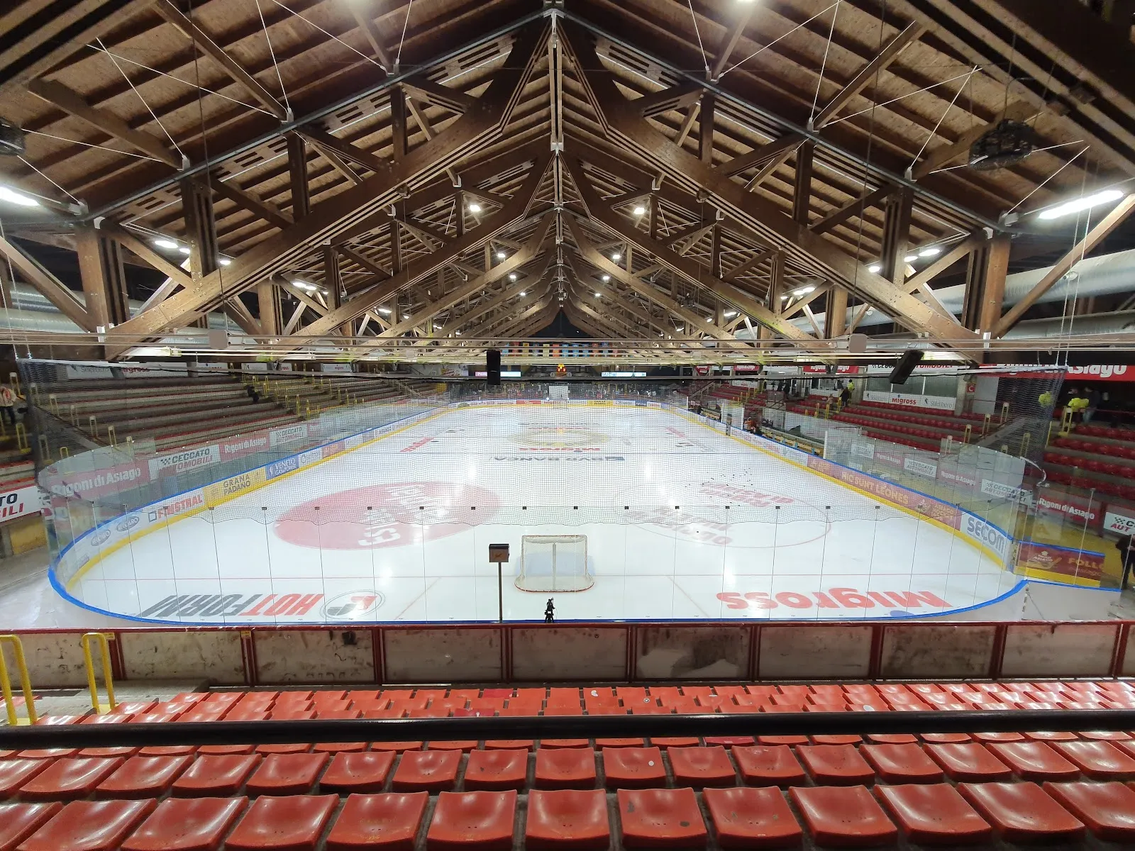 The ice of the Pala Hodegart seen from the red seats, under the laminated-timber roof, with the boards and goal at the far end. — Pala Hodegart