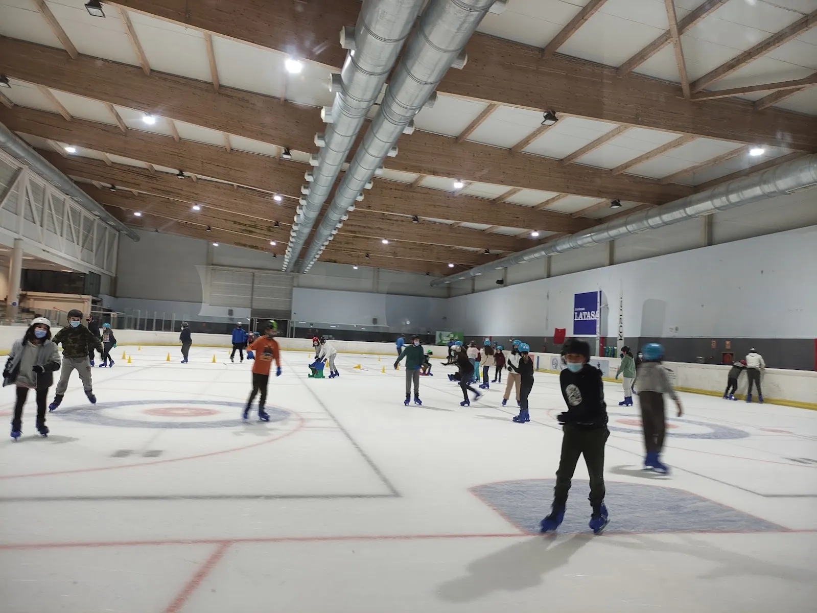 The wide interior of the ice hall: the full rink with skaters small at the far side, the roof trusses and the ducts above. — Palacio de Hielo de Huarte