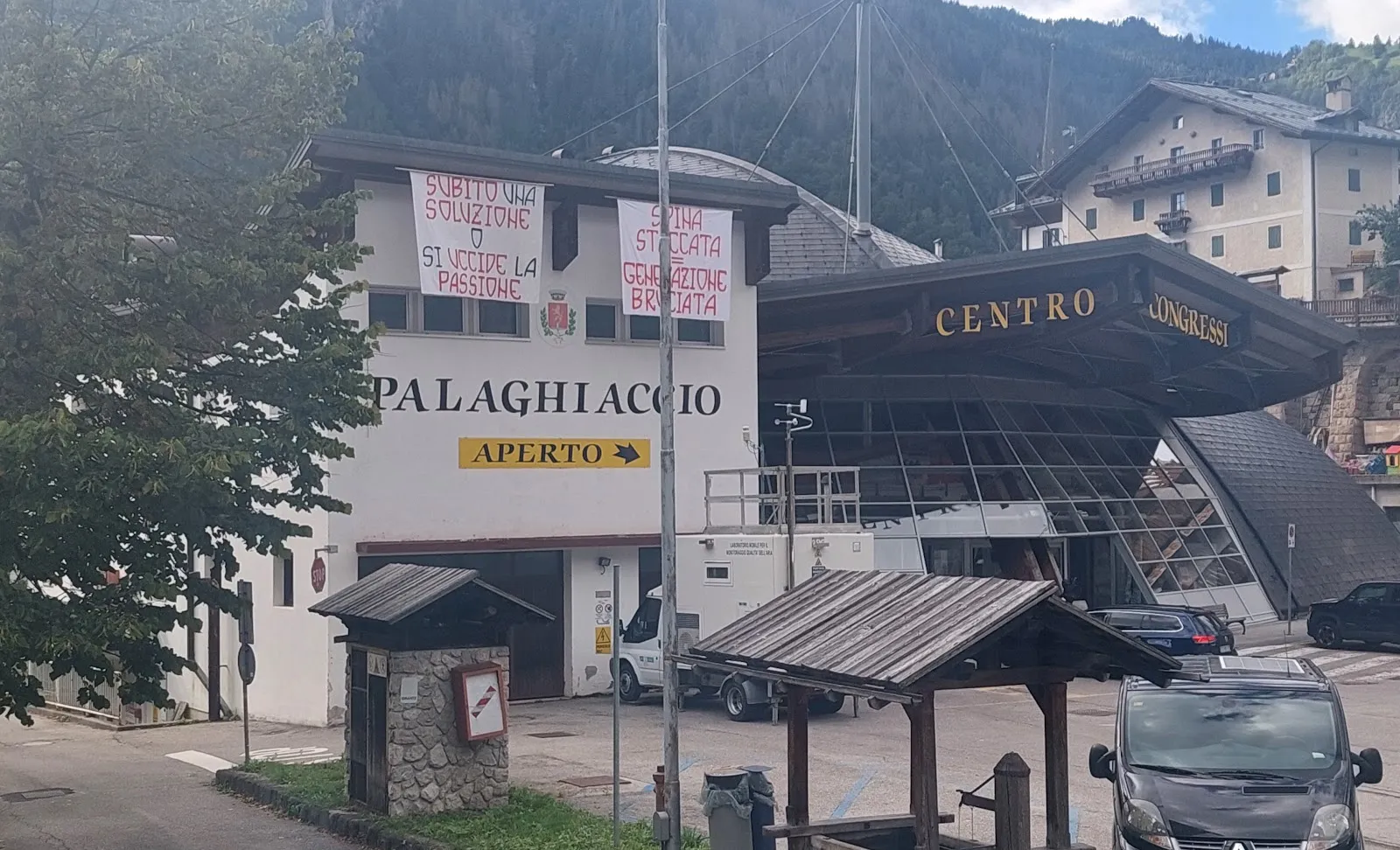 The Alleghe Palaghiaccio building with its sign above the entrance and the mountains rising behind the lake road. — Palaghiaccio Alvise De Toni (Alleghe)