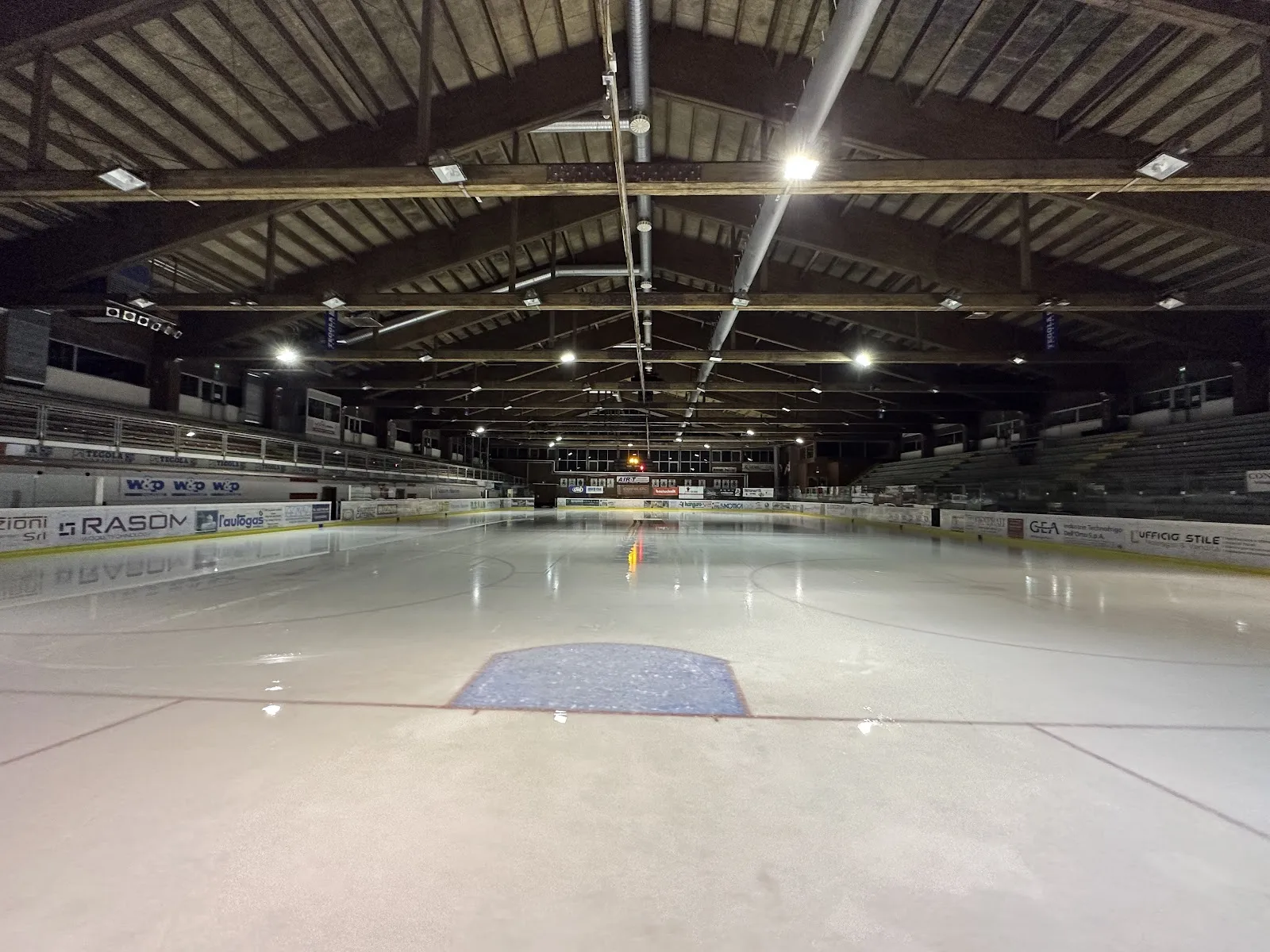 The empty ice under the timber roof of the Alleghe palaghiaccio, with the oval boards and a blue centre mark. — Palaghiaccio Alvise De Toni (Alleghe)