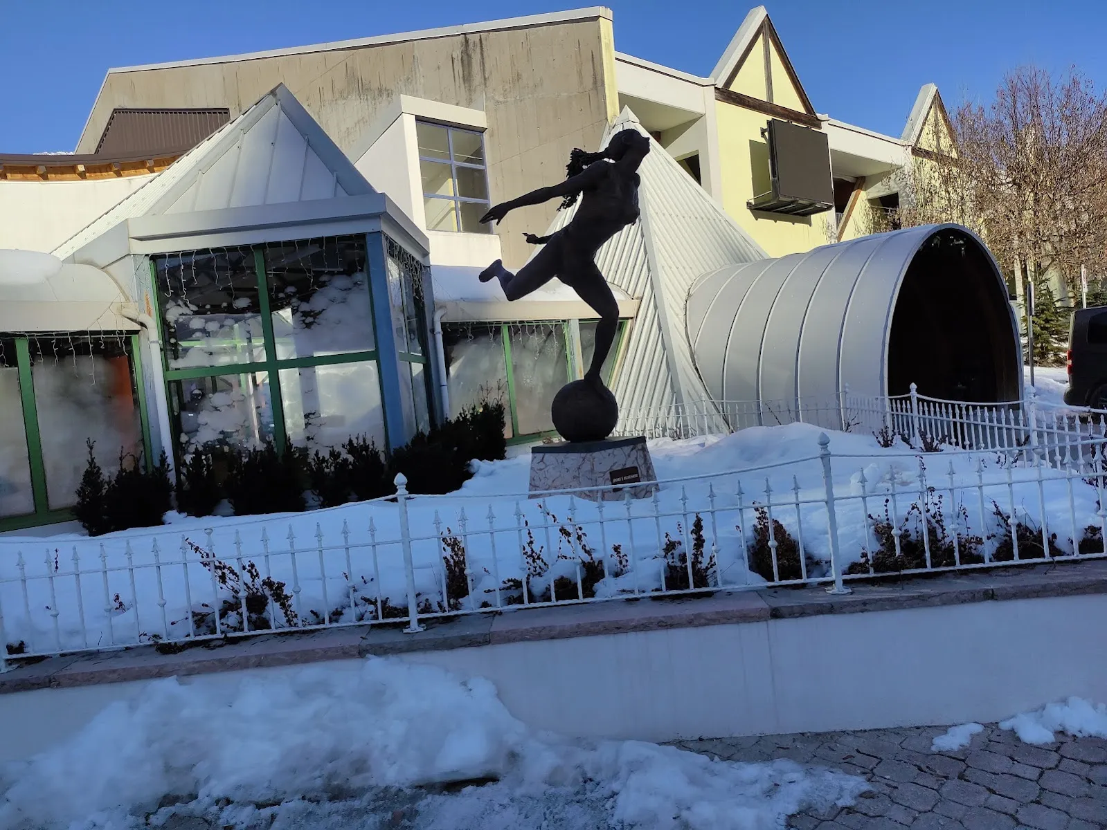 The snow-covered outside of the Andalo Life sports complex that houses the rink, with a glazed pavilion, an arched passage and a statue of a footballer by the fence. — Palaghiaccio Andalo