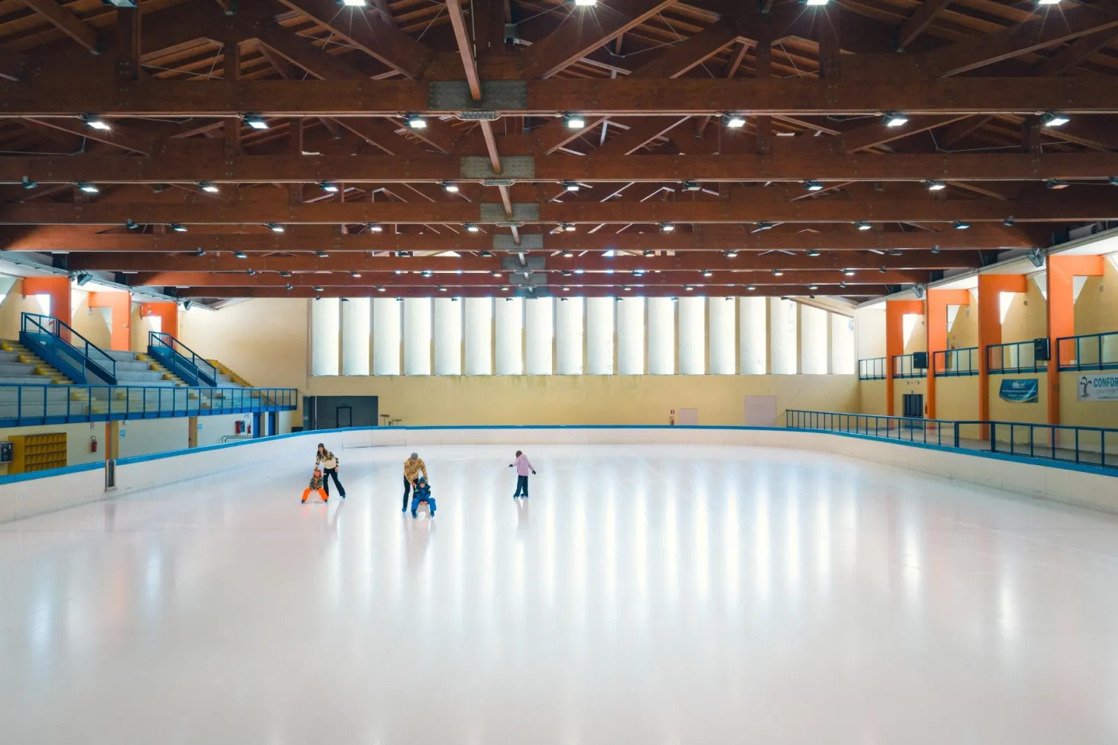 The wide ice sheet of the covered hall under its timber roof, with a few skaters far down the rink and the boards and stands along the sides. — Palaghiaccio Andalo