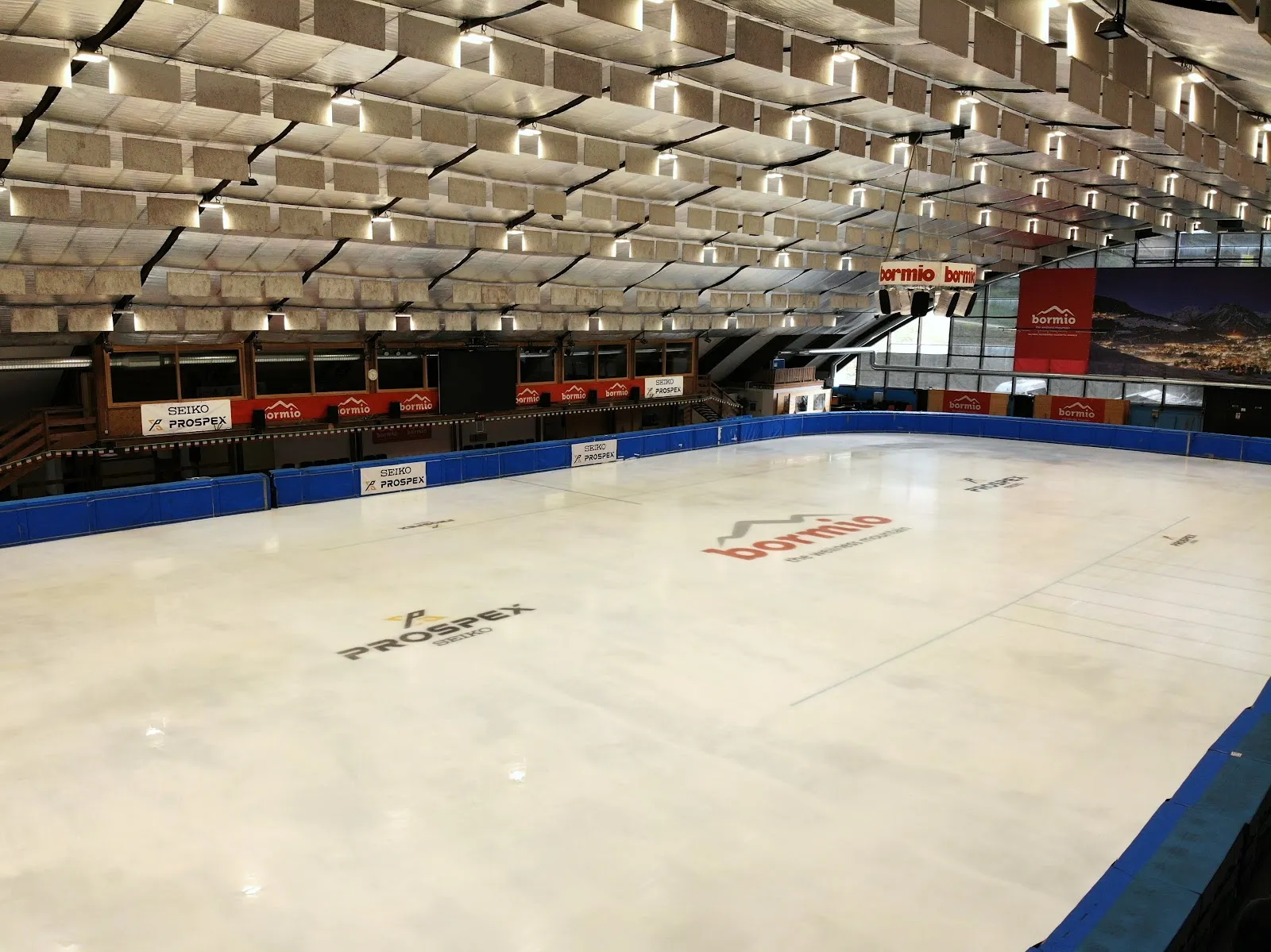 The empty ice sheet of the Bormio palaghiaccio seen from the side, with blue boards, the orange seating and the sound-absorbing roof panels above. — Palaghiaccio Bormio (Palaghiaccio Braulio)