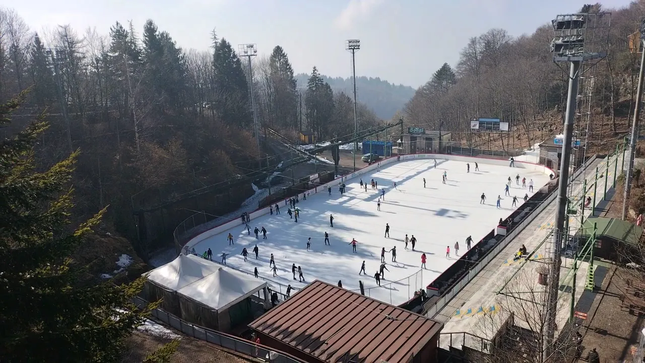 Aerial view of the open-air ice rink at Bosco Chiesanuova in the woods, with skaters on the sheet and the entrance huts — Palaghiaccio di Bosco Chiesanuova