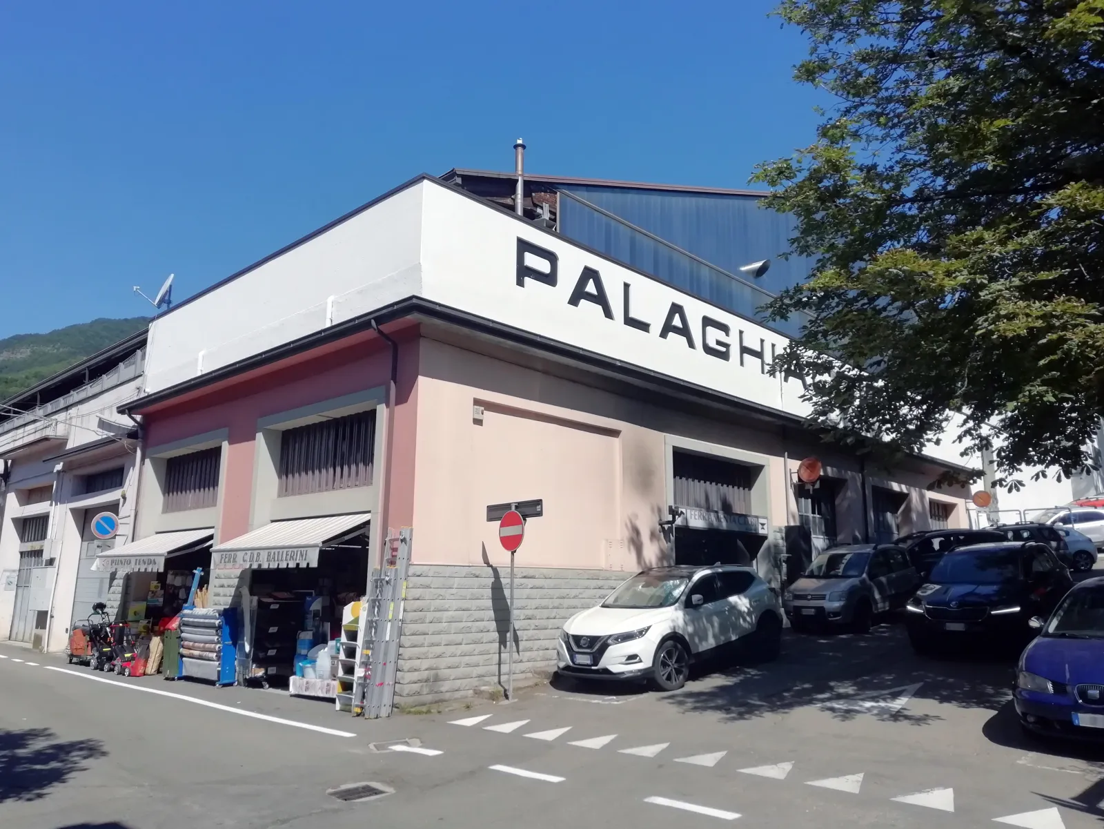 The corner of the Palaghiaccio building in Fanano seen from the street, with the name across the upper wall, shops at street level and parked cars — Palaghiaccio Fanano