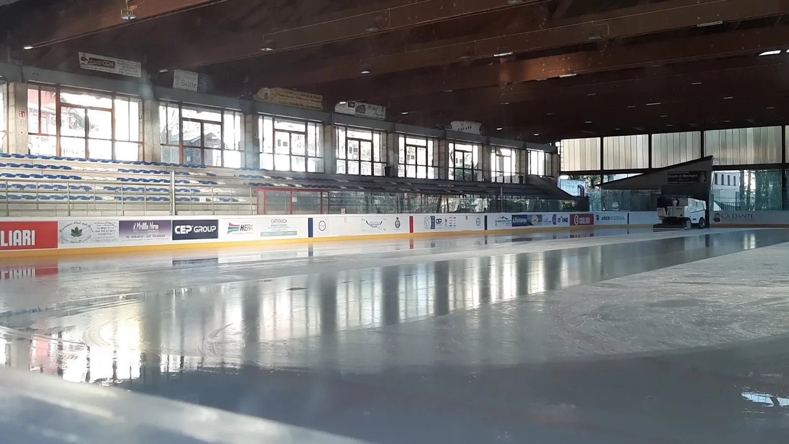 The empty ice sheet seen from beside the boards, with the wooden roof, the glazed upper wall and the stand behind. — Palaghiaccio Fanano