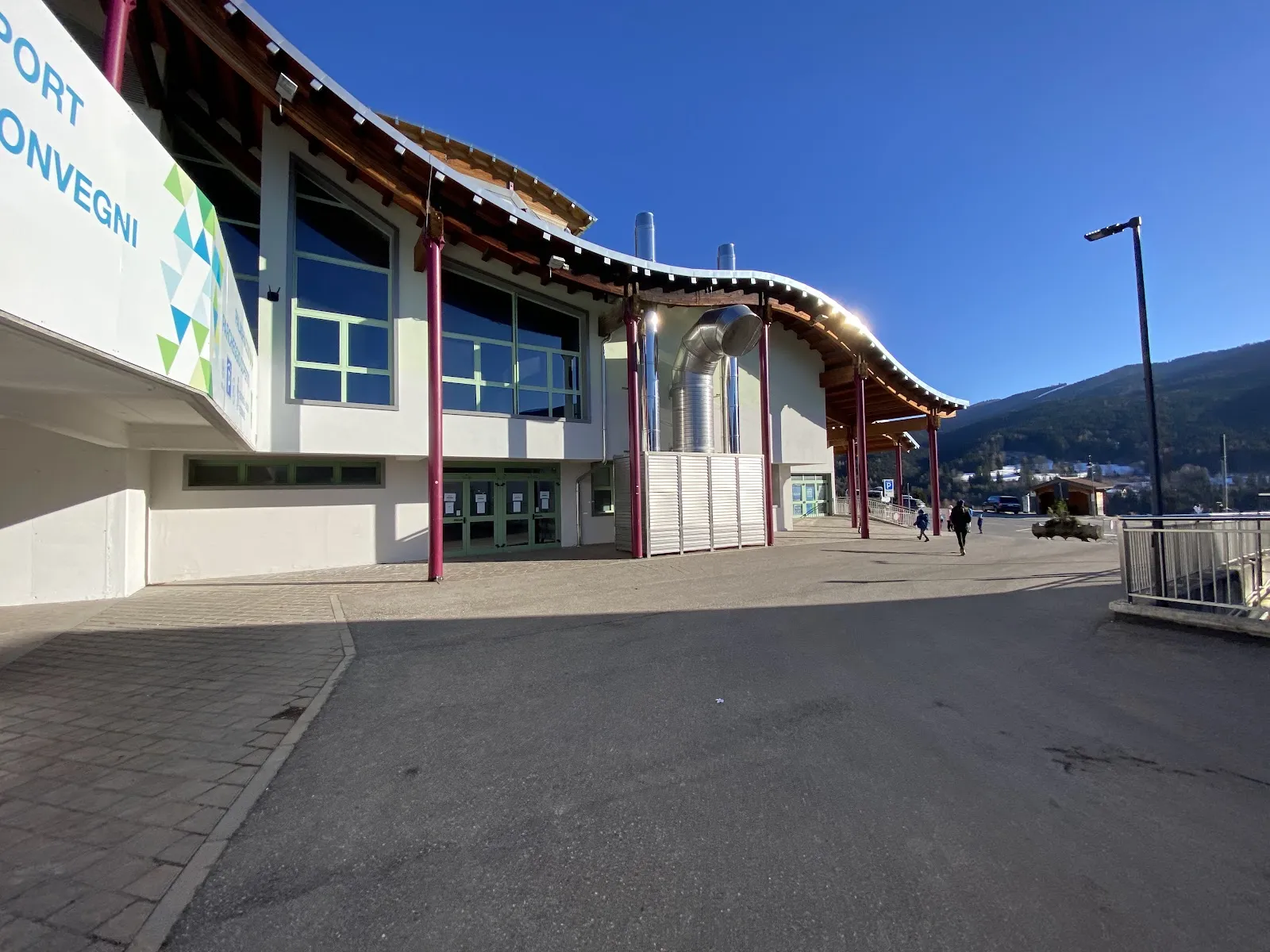 The curved timber-roofed front of the PalaFolgaria sports and convention hall in Folgaria, seen from the forecourt on a sunny day. — Palaghiaccio Folgaria