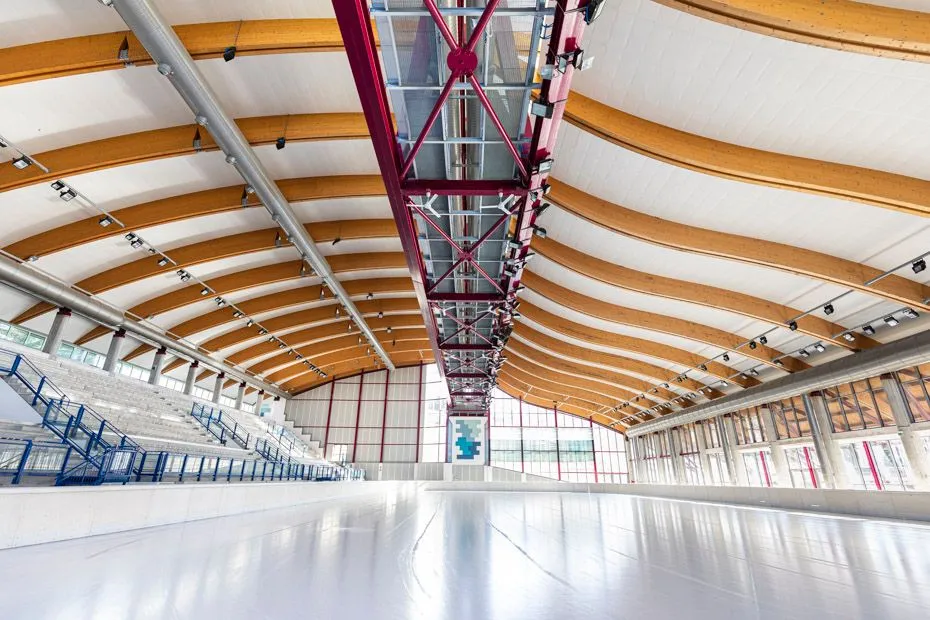 The empty ice sheet of the covered hall seen from one end, under the curved timber roof beams, with the stand and the glazed side wall along the rink. — Palaghiaccio Folgaria