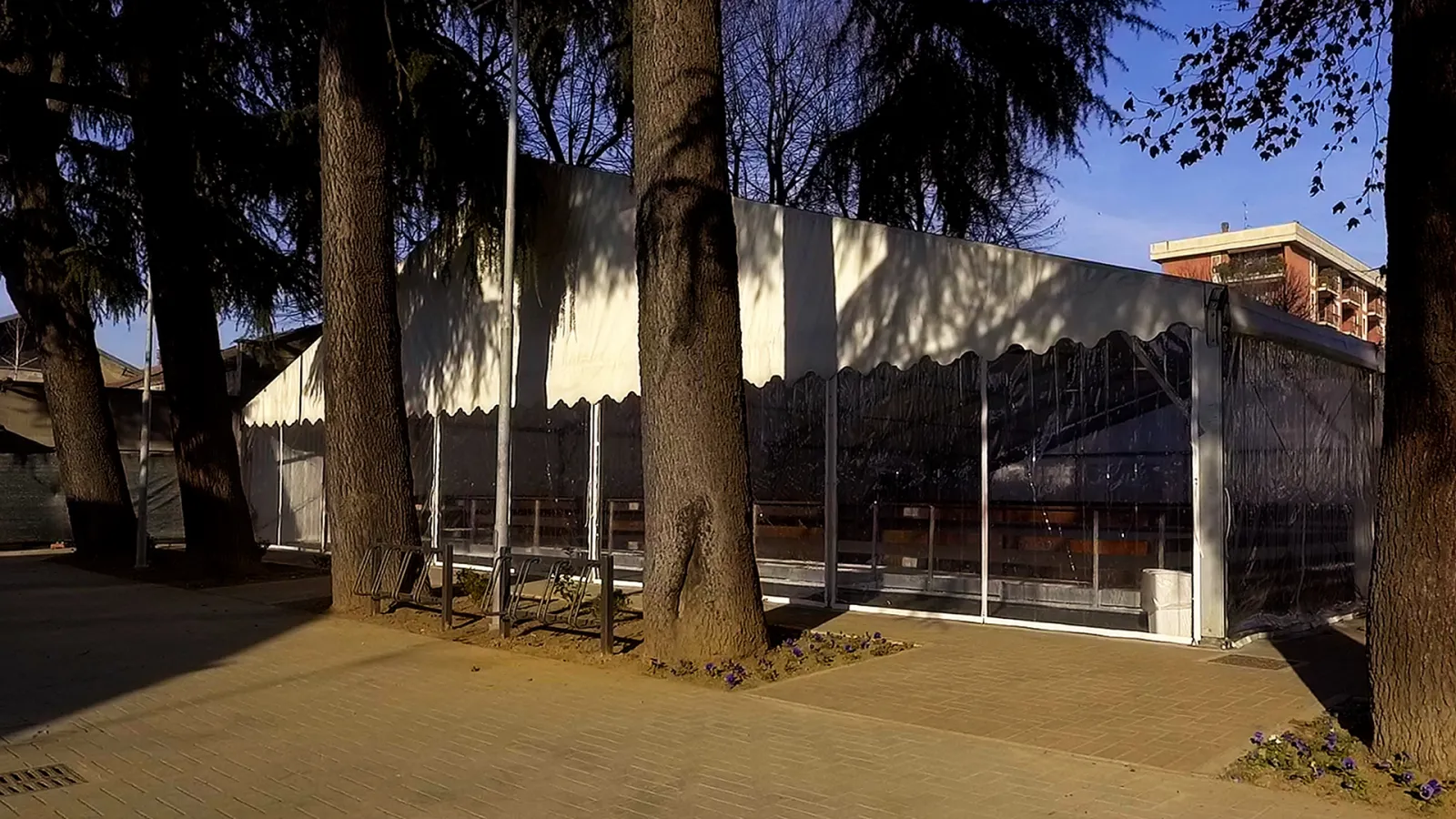 The white tent hall of the ice rink between the trees on via Piave, seen from the path outside. — Palaghiaccio PalaExbo Saronno