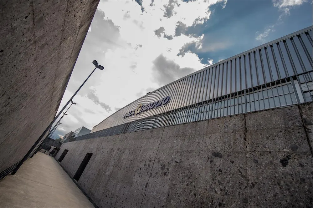 The grey metal-clad side of the PalaGhiaccio building in Trento with its name sign, seen from below a concrete wall. — PalaGhiaccio Trento (Maccani Ice Arena)