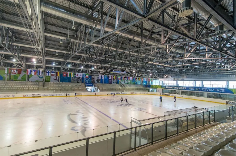 The 60 by 30 metre ice of the PalaGhiaccio in Trento seen from the stands under the steel roof trusses, with a few skaters on the ice and the Winter Universiade 2013 banners along the boards. — PalaGhiaccio Trento (Maccani Ice Arena)