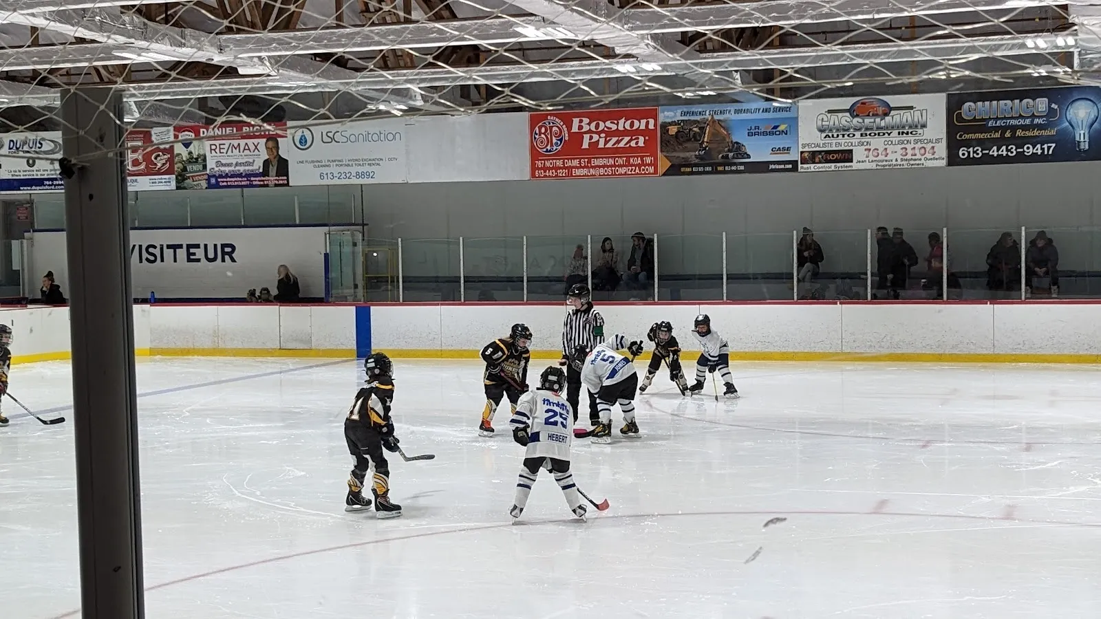 Un saque de hockey juvenil en el hielo del Palais des Sports d'Embrun, con la banca de los visitantes y los espectadores a lo largo del lado opuesto, bajo una hilera de vallas de patrocinadores locales. — Palais des Sports (Embrun Arena)