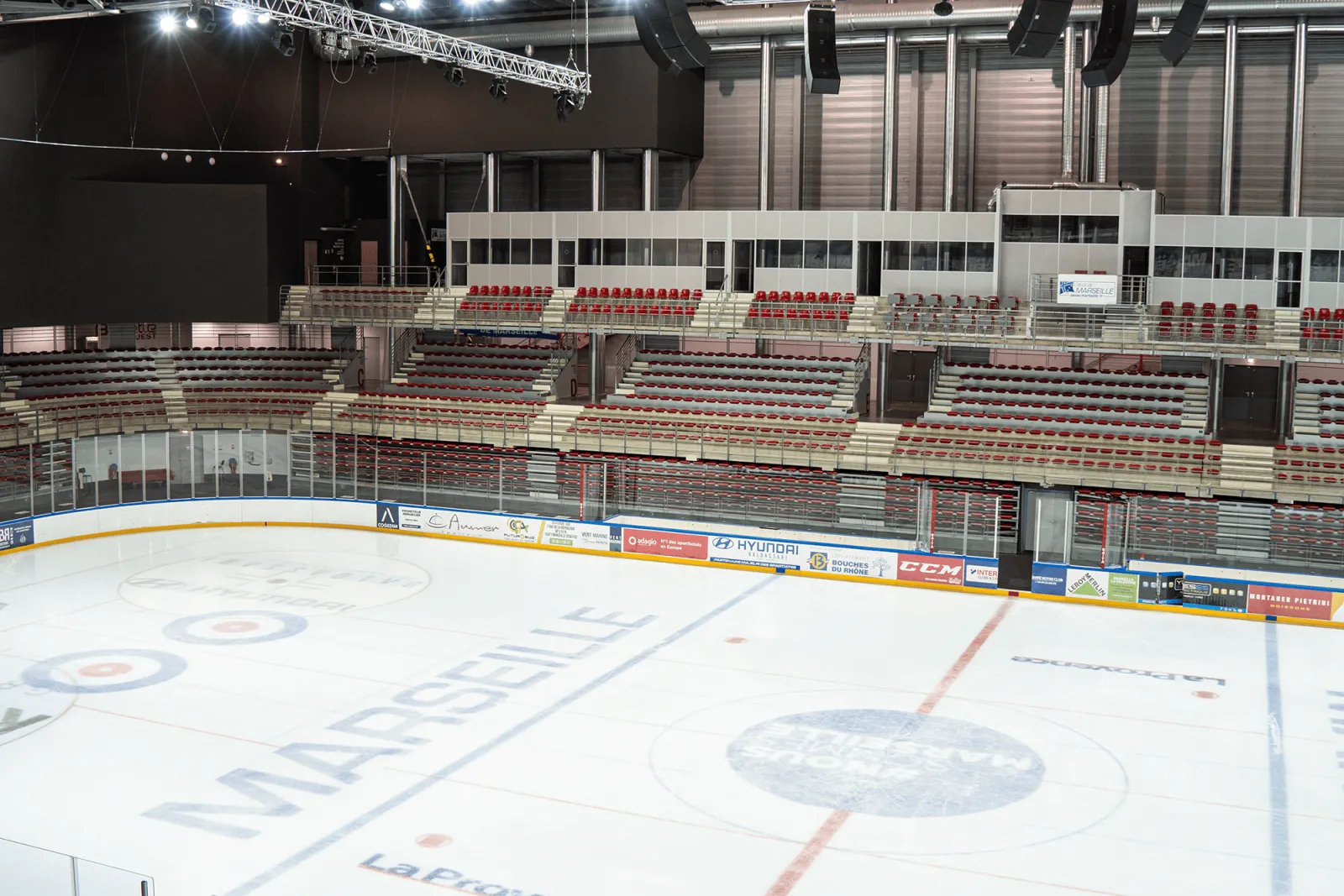 The Olympic ice sheet seen from the boards, with MARSEILLE lettered on the ice beside curling rings and faceoff circles, and tiers of red and grey stands behind sponsor boards. — Patinoires du Palais omnisports Marseille Grand-Est