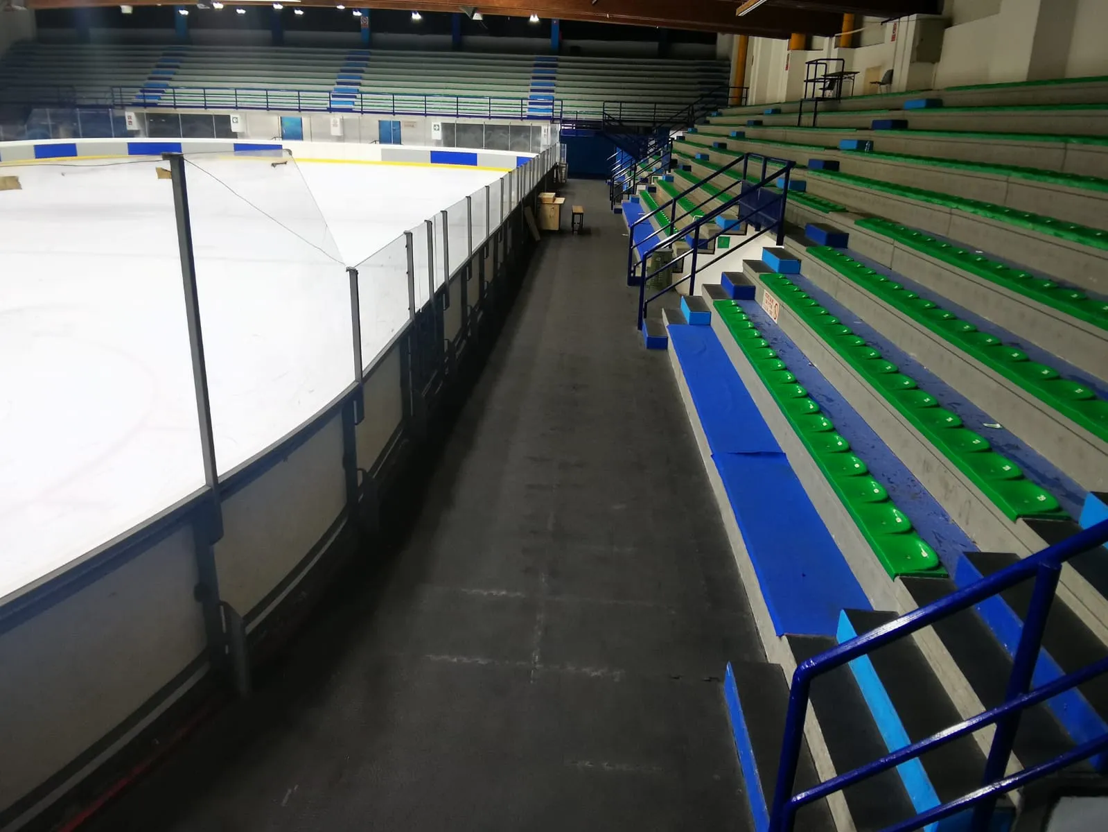 An empty ice sheet at the PalaSesto seen from the stands, with green and blue bench seating along the boards. — PalaSesto